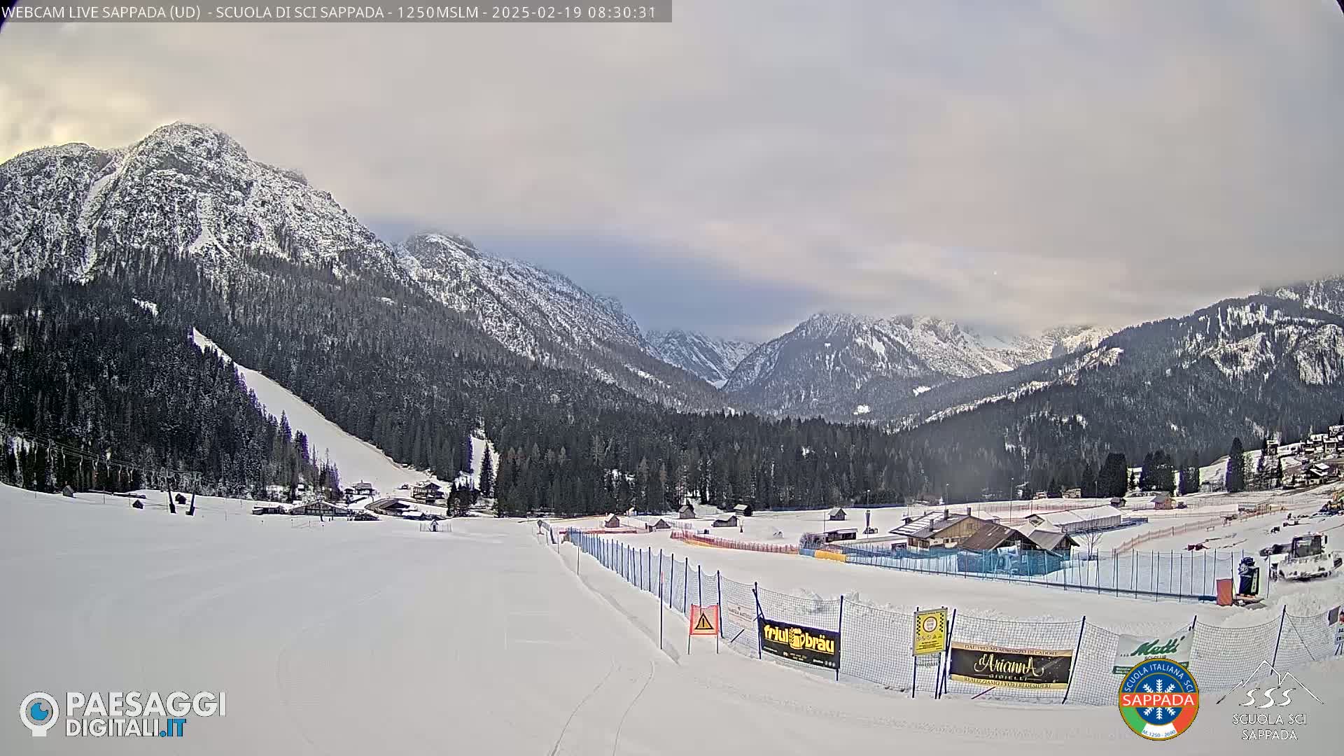A snow-covered ski slope leads to a valley nestled between snow-dusted mountains under an overcast sky.