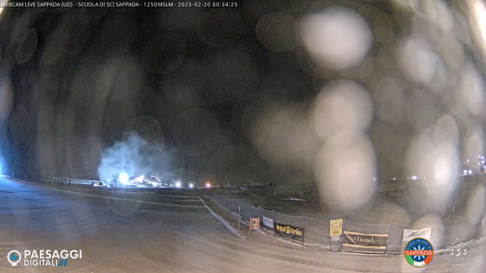 A nighttime snow scene with a hazy atmosphere shows lights and snow-covered ground at a ski resort.