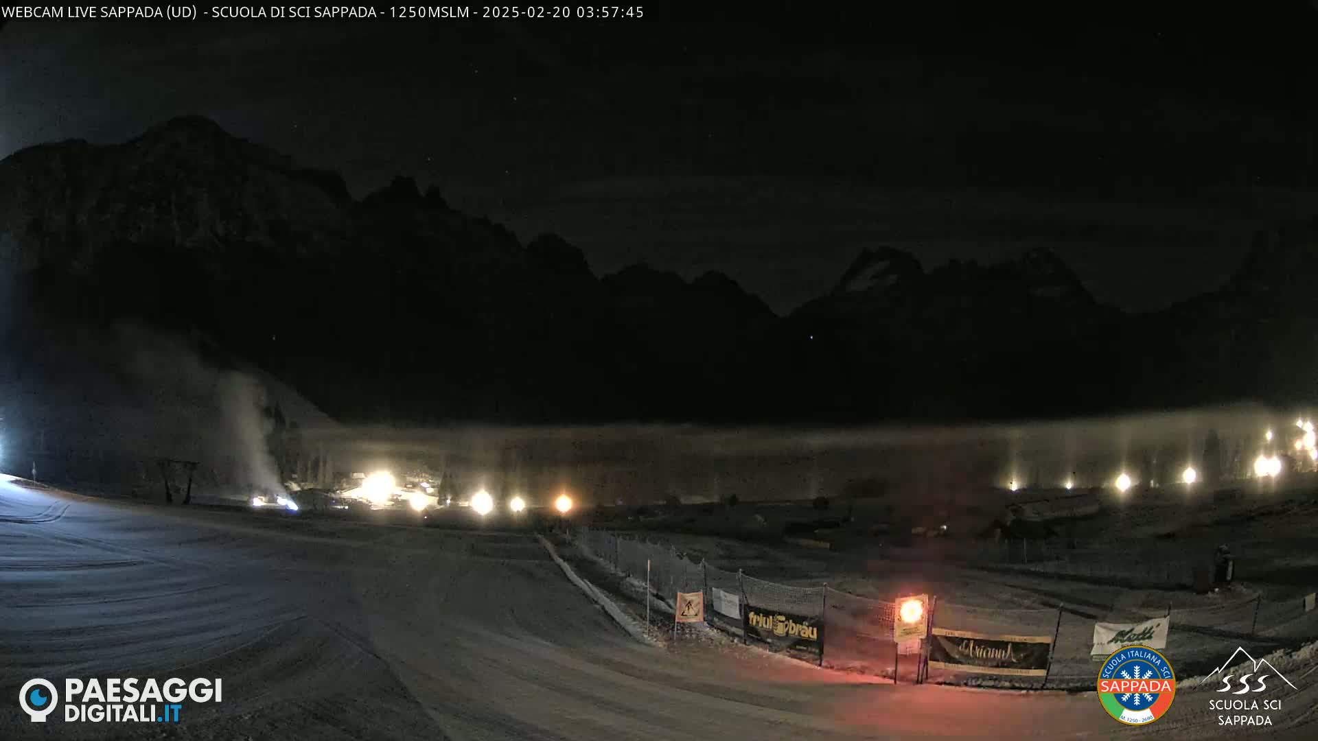 A snow-covered ski slope at night is illuminated by several lights against a backdrop of dark mountains under a mostly clear sky.