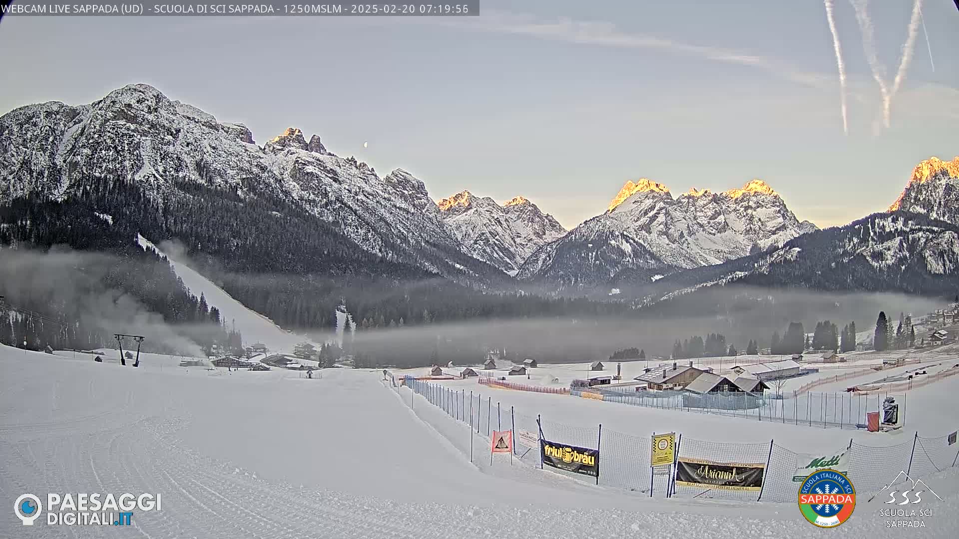 A snow-covered ski slope and valley are nestled beneath a range of snow-capped mountains, with a light fog settling in the valley and the sun beginning to rise, illuminating the peaks.