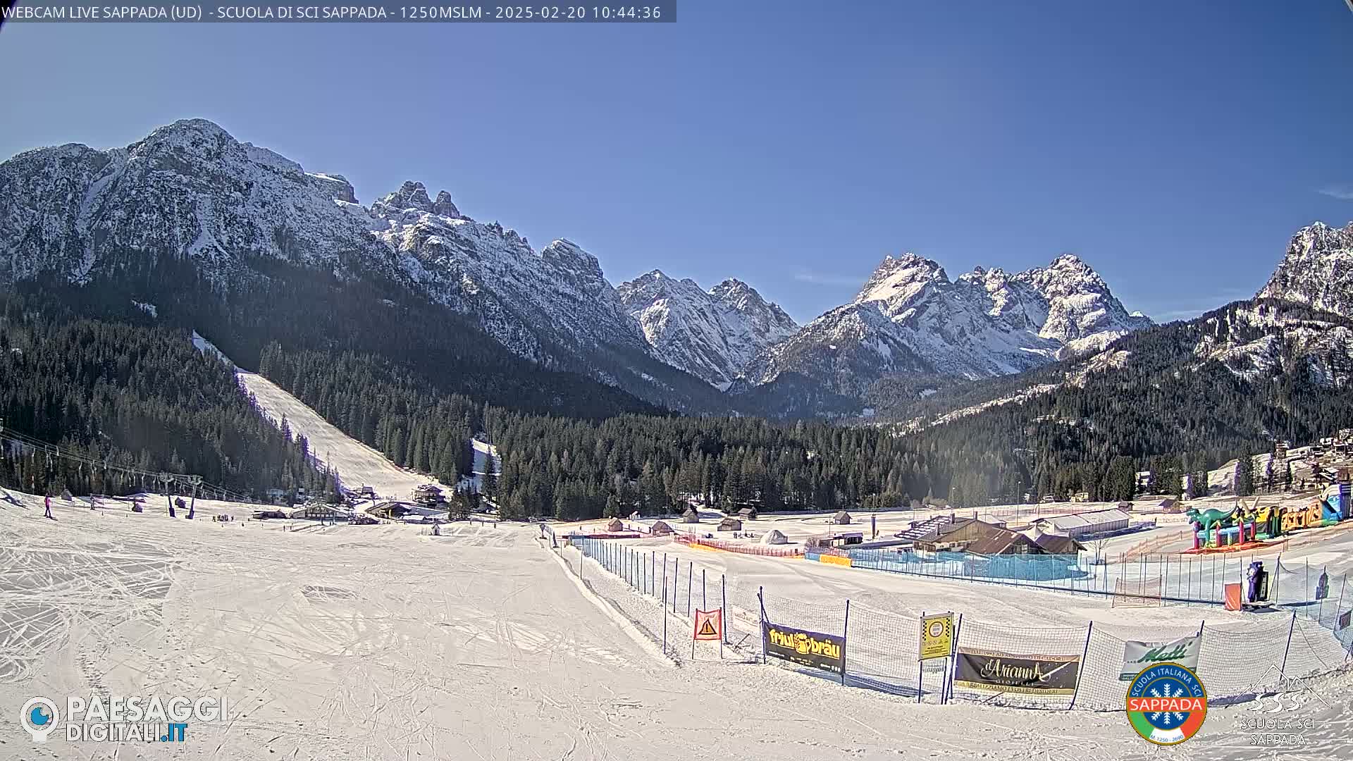 A snow-covered ski slope is nestled in a valley, with snow-capped mountains rising in the background under a clear blue sky.