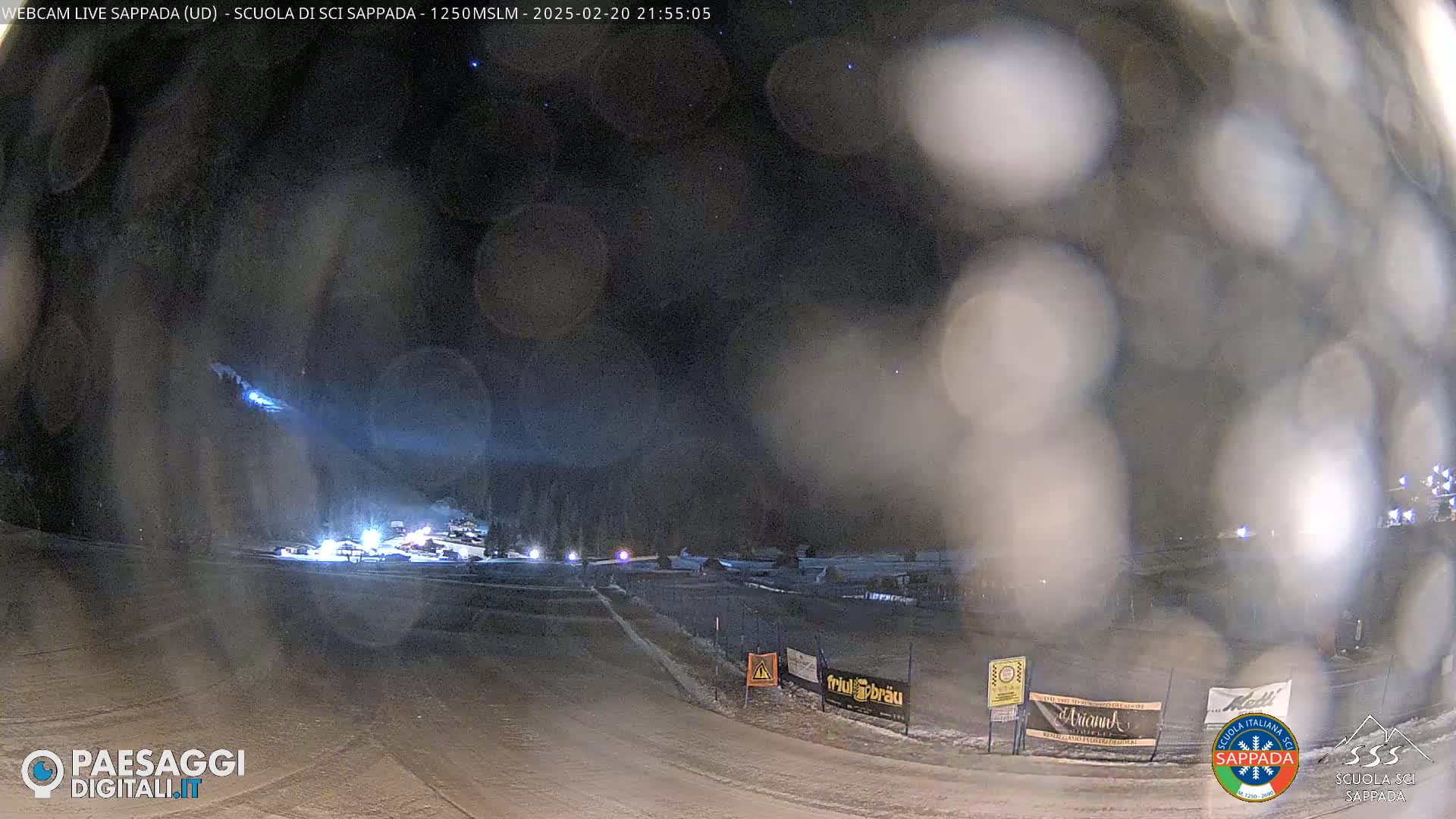 A nighttime view of a snow-covered ski resort with lights illuminating a road and buildings under a dark, starry sky with some light snow or fog obscuring the view.