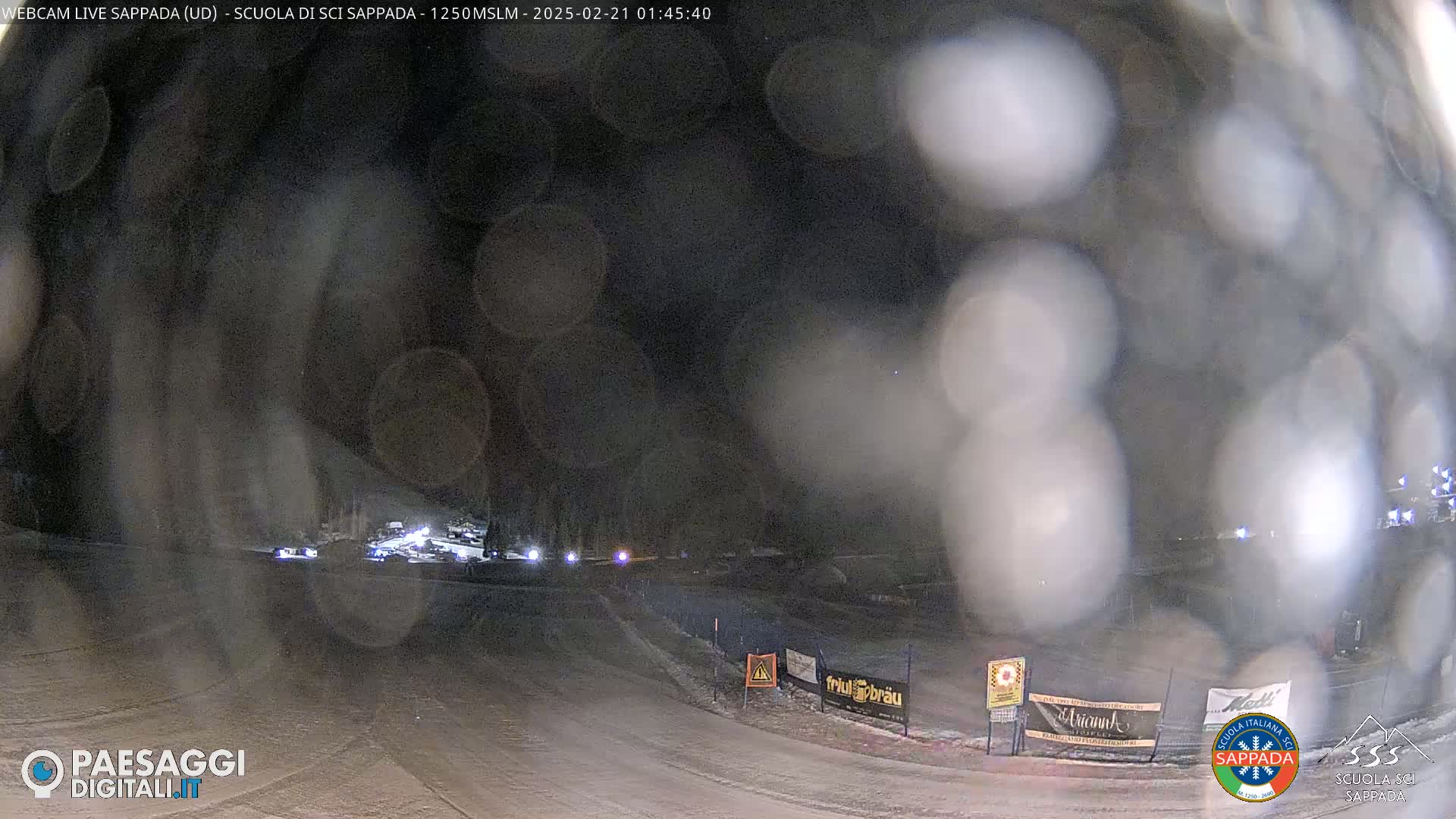 A nighttime, snowy scene shows a road with a few vehicles and some signage, viewed through a blurry, possibly rain- or snow-covered lens.