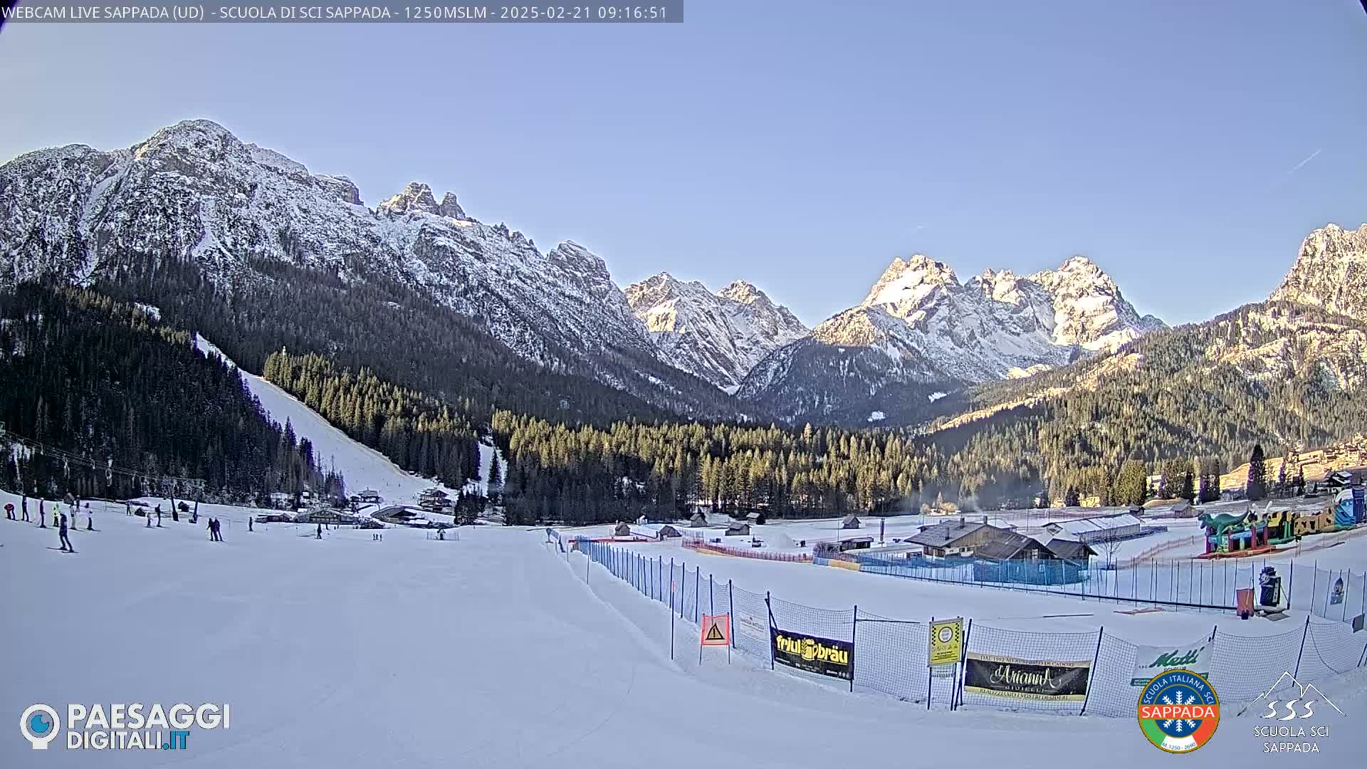 A snow-covered ski slope sits below snow-capped mountains under a clear blue sky.