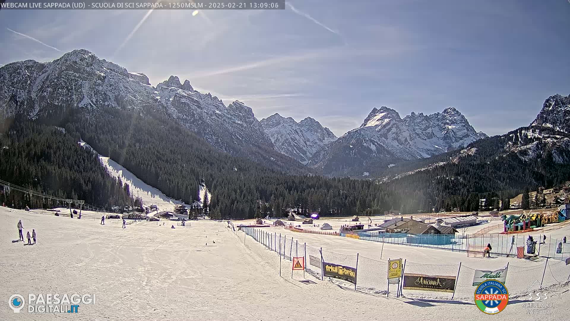 A snow-covered ski slope with a few skiers, nestled in a valley surrounded by snow-capped mountains under a mostly sunny sky.