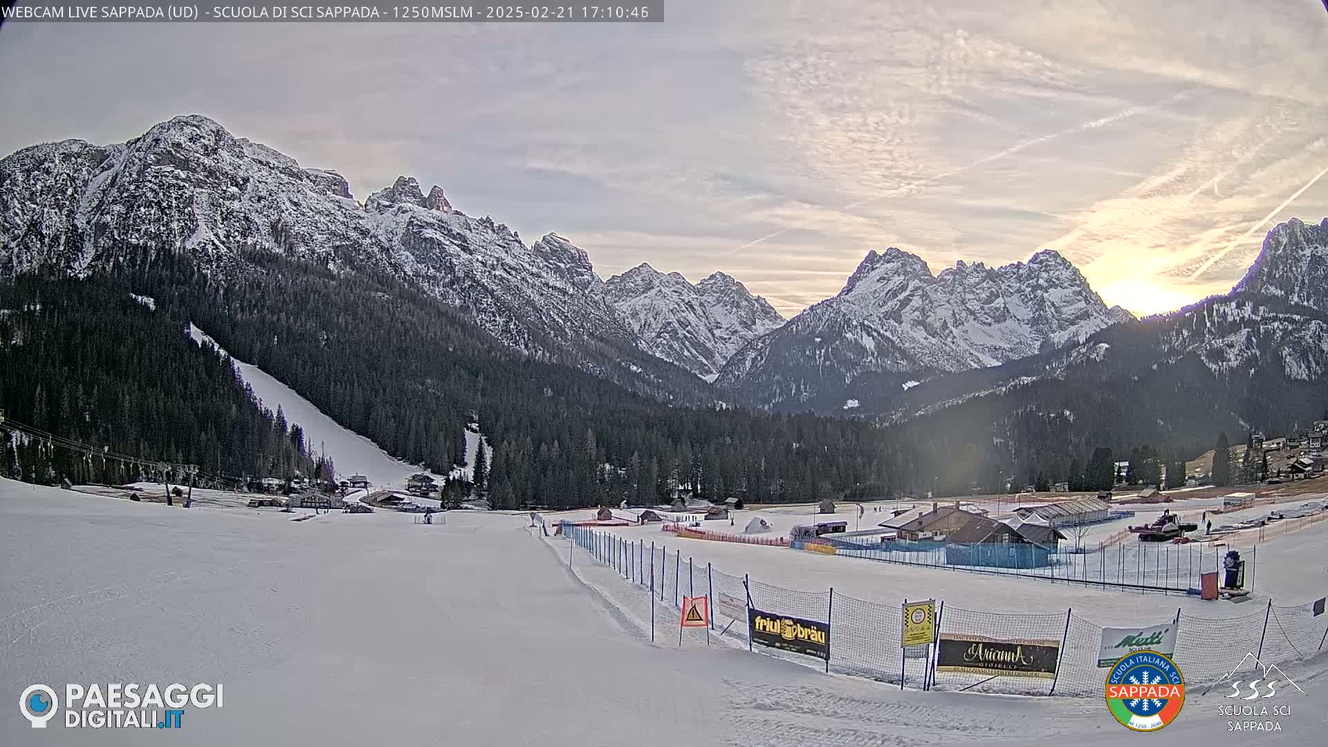 A snow-covered ski slope and valley are nestled beneath a range of snow-capped mountains, under a partly cloudy sky with a sunset.