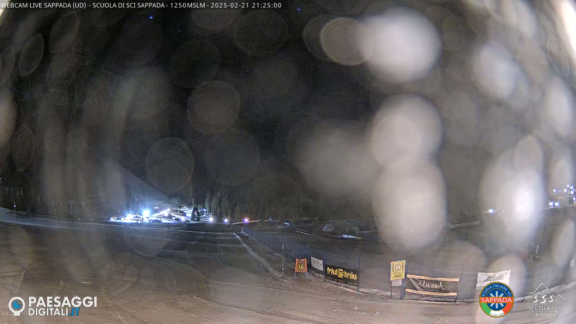 A nighttime view of a snow-covered ski area with several lights visible, under a mostly clear sky with a few stars.