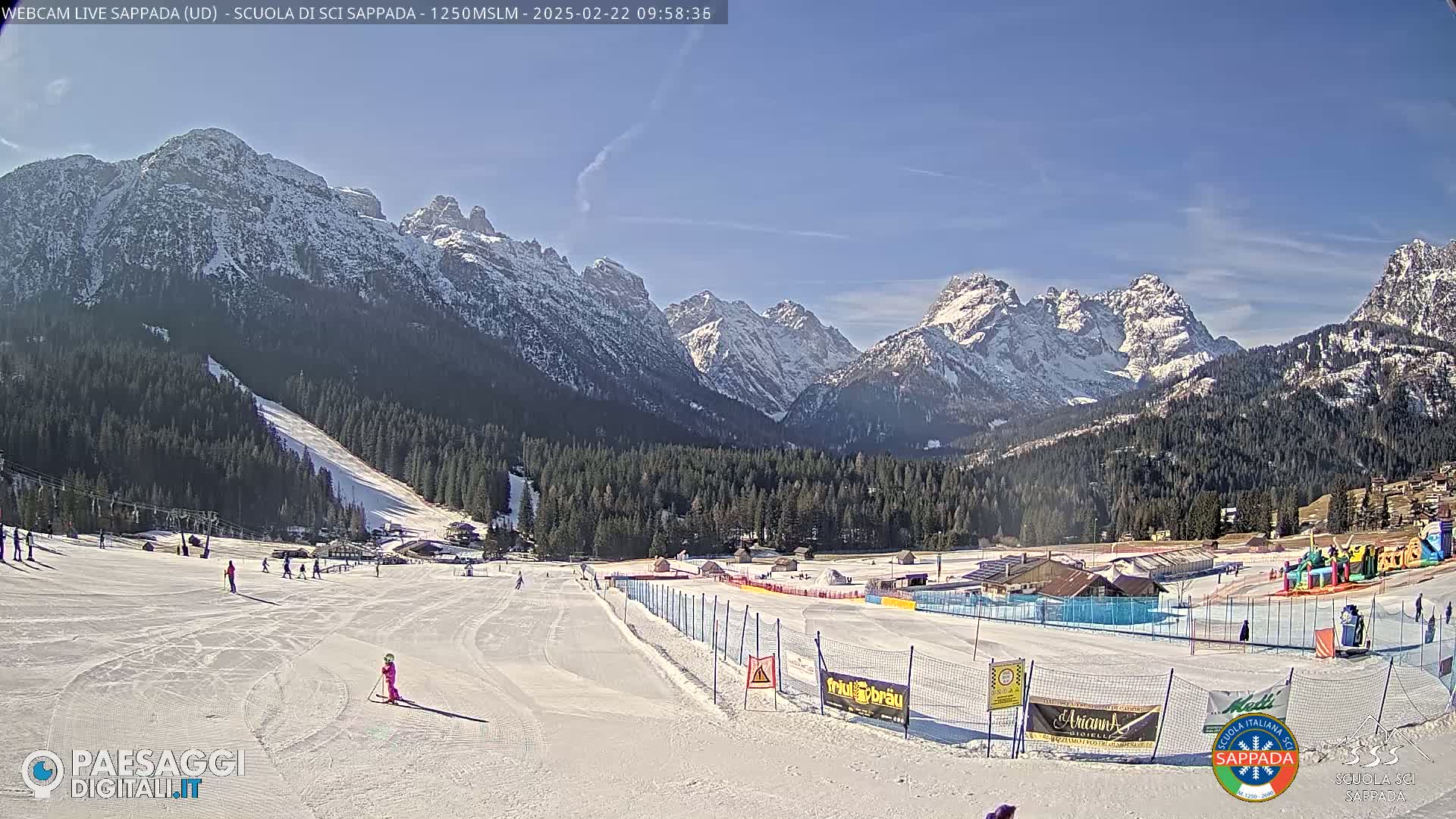 A sunny day reveals a snow-covered ski slope with several skiers, surrounded by a snow-capped mountain range and a coniferous forest.