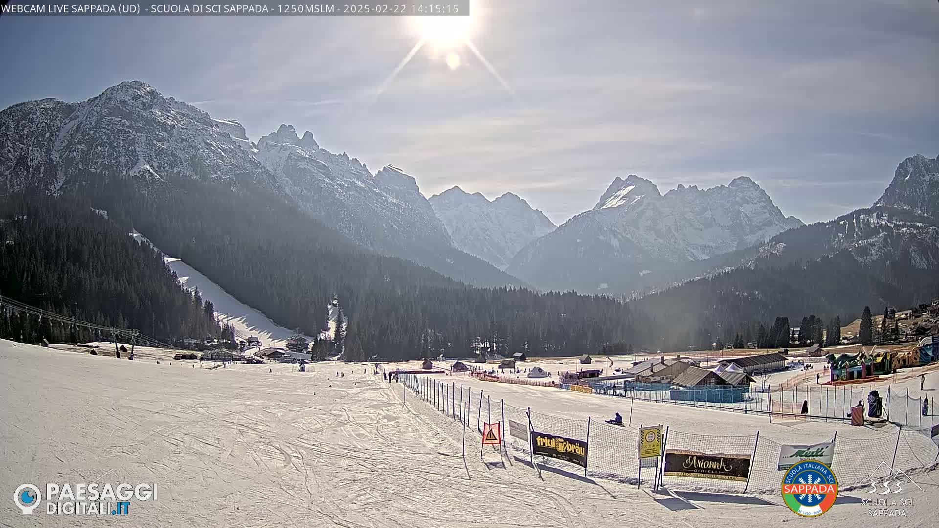 A sunny day reveals a snow-covered ski slope at the foot of snow-capped mountains, with several buildings and people visible in the distance.