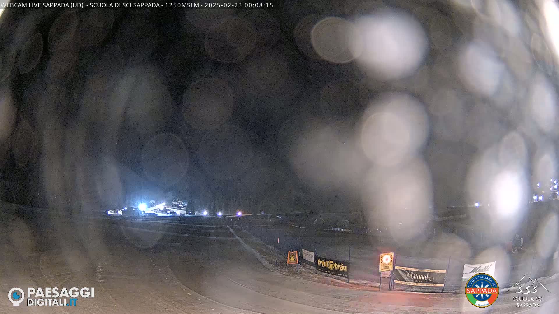 A nighttime view of a snow-covered area with lights in the distance, obscured by blurry, possibly snowy or foggy conditions.