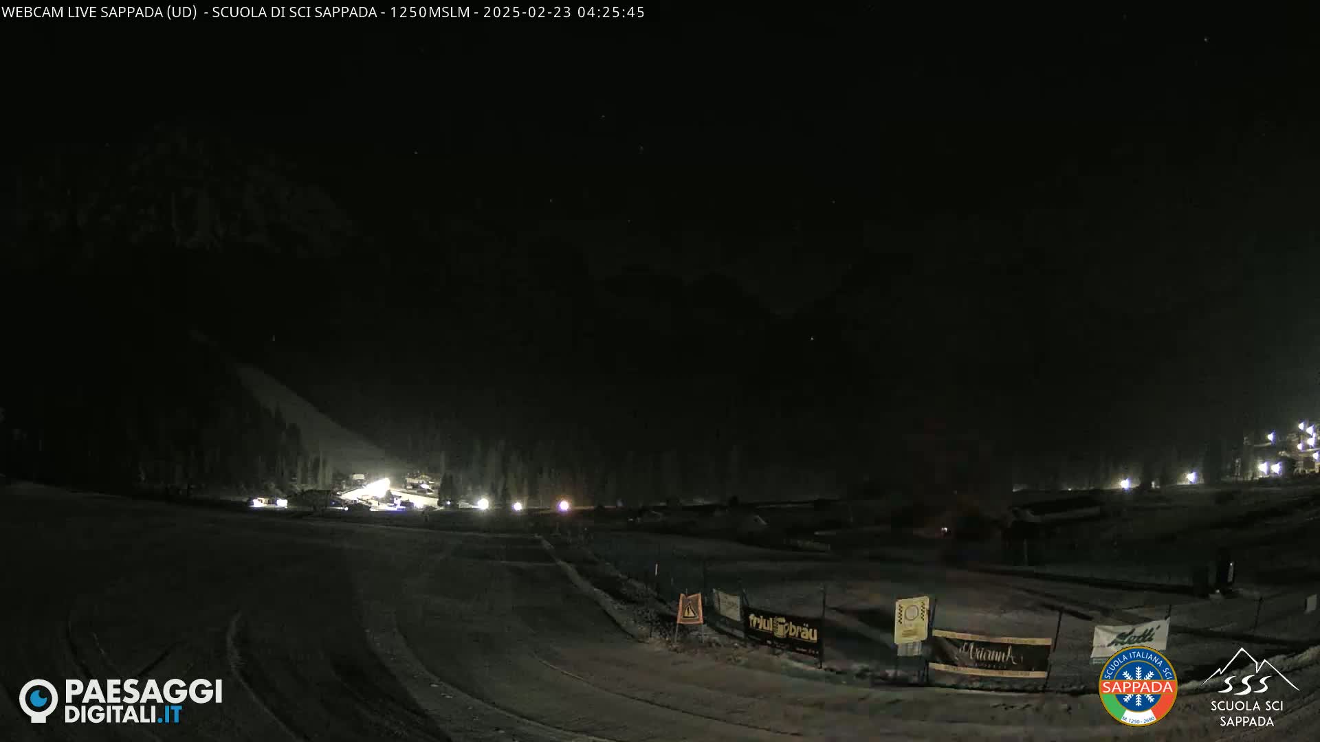 A nighttime view of a snow-covered ski slope with a few lights visible in the distance under a clear, dark sky.