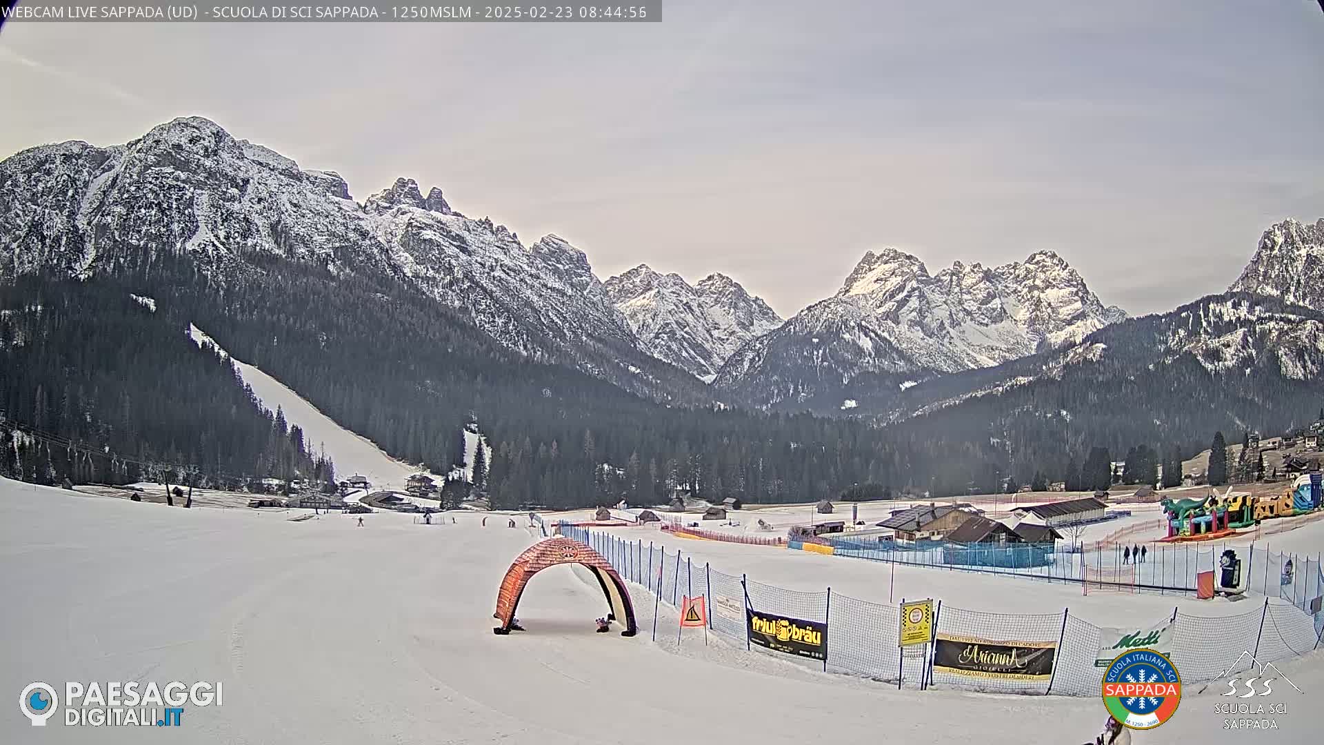 A snow-covered ski slope with a mountain range in the background under a mostly cloudy sky.