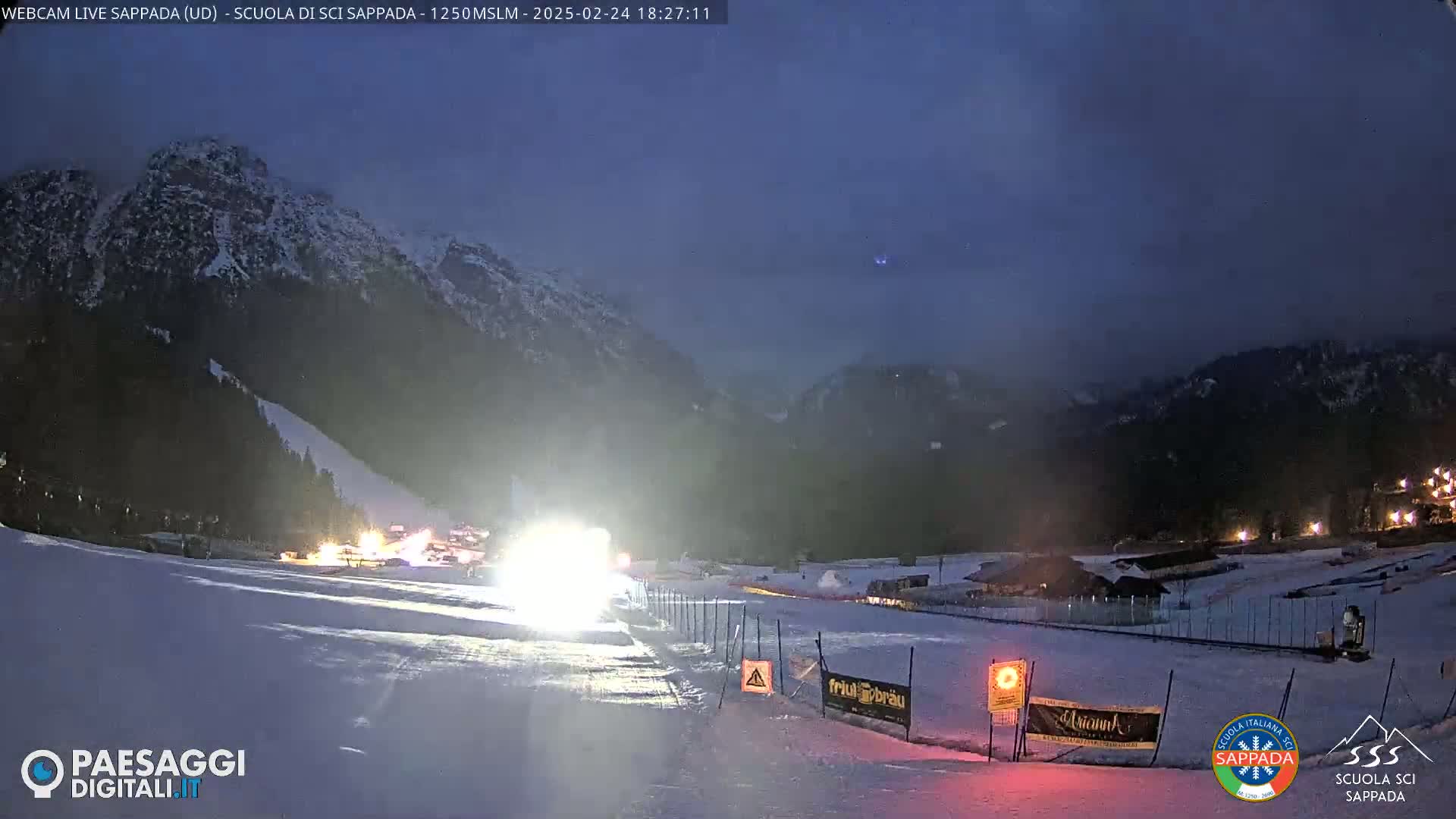 A nighttime view of a snow-covered ski slope, illuminated by headlights, with mountains in the background under a dark, hazy sky.