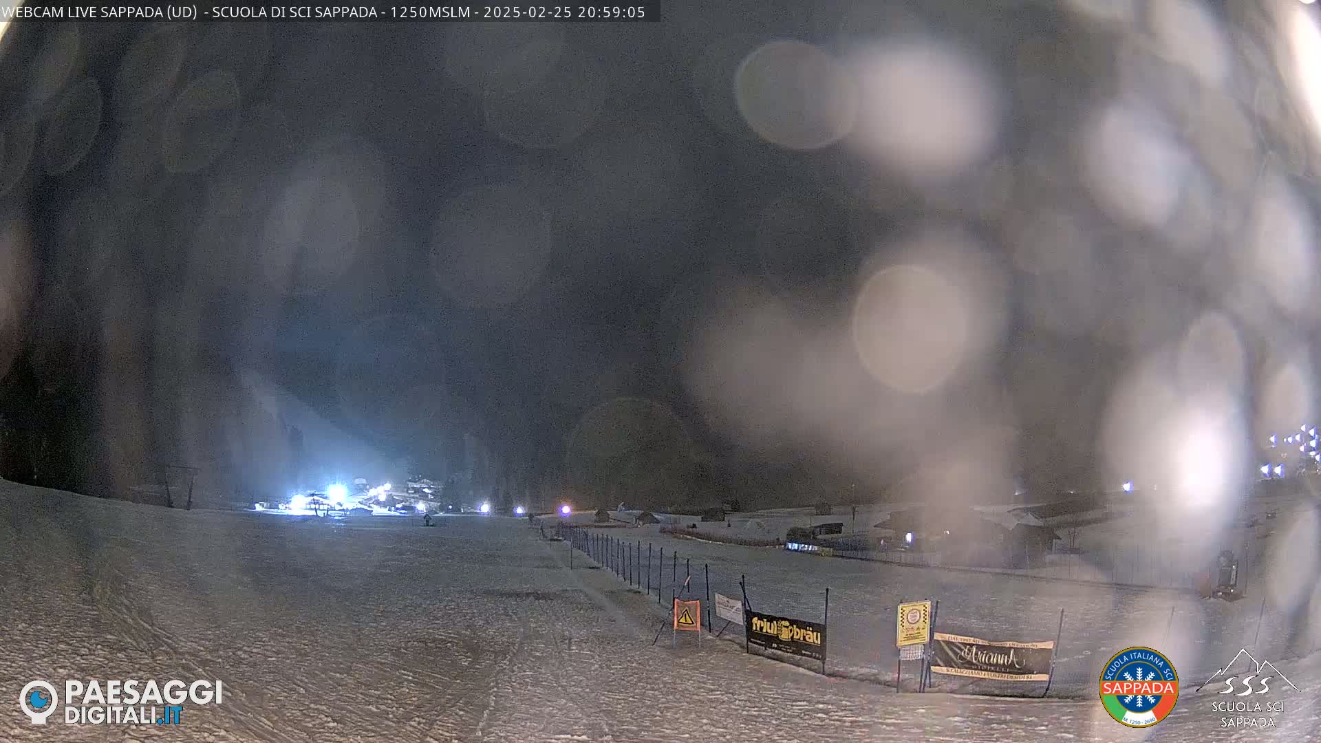A snow-covered ski slope at night, dimly lit with some lights in the distance, under a dark, possibly snowy sky.