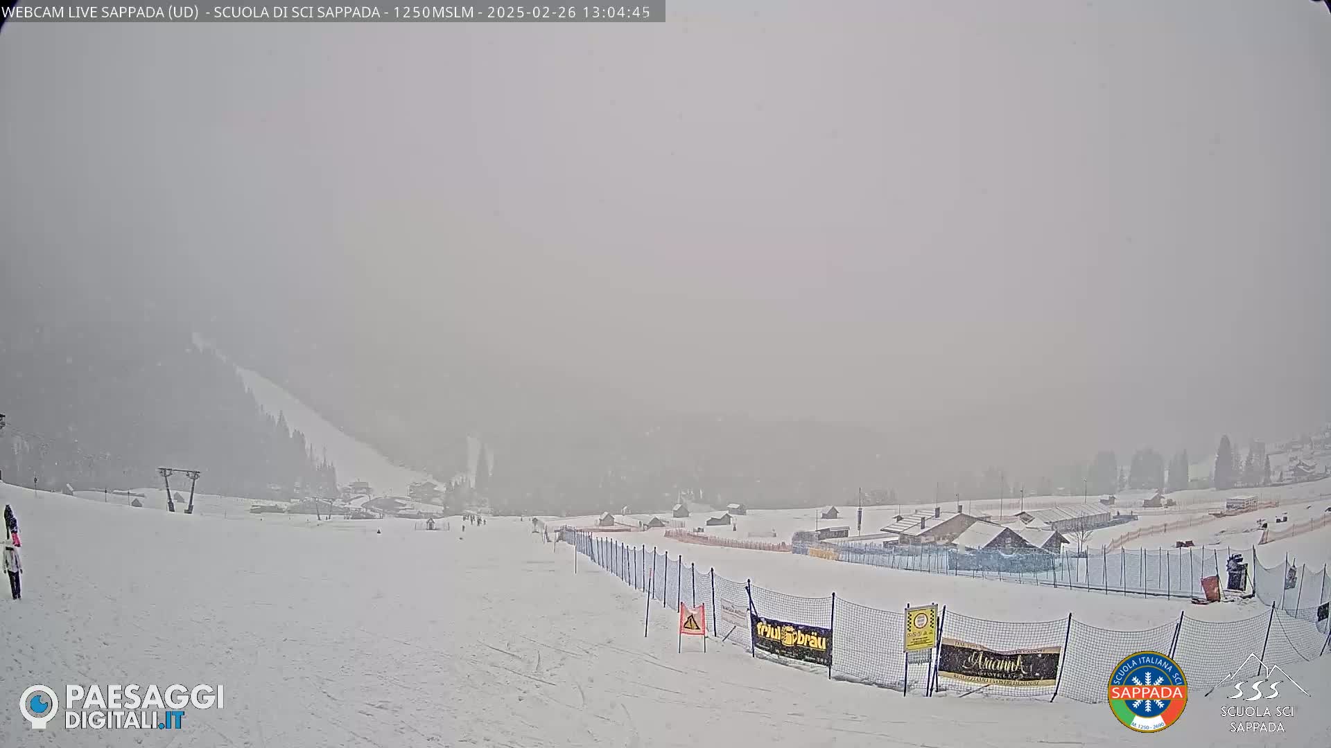 A snowy, foggy scene of a ski slope with a few people visible in the distance and a fence running down the middle.