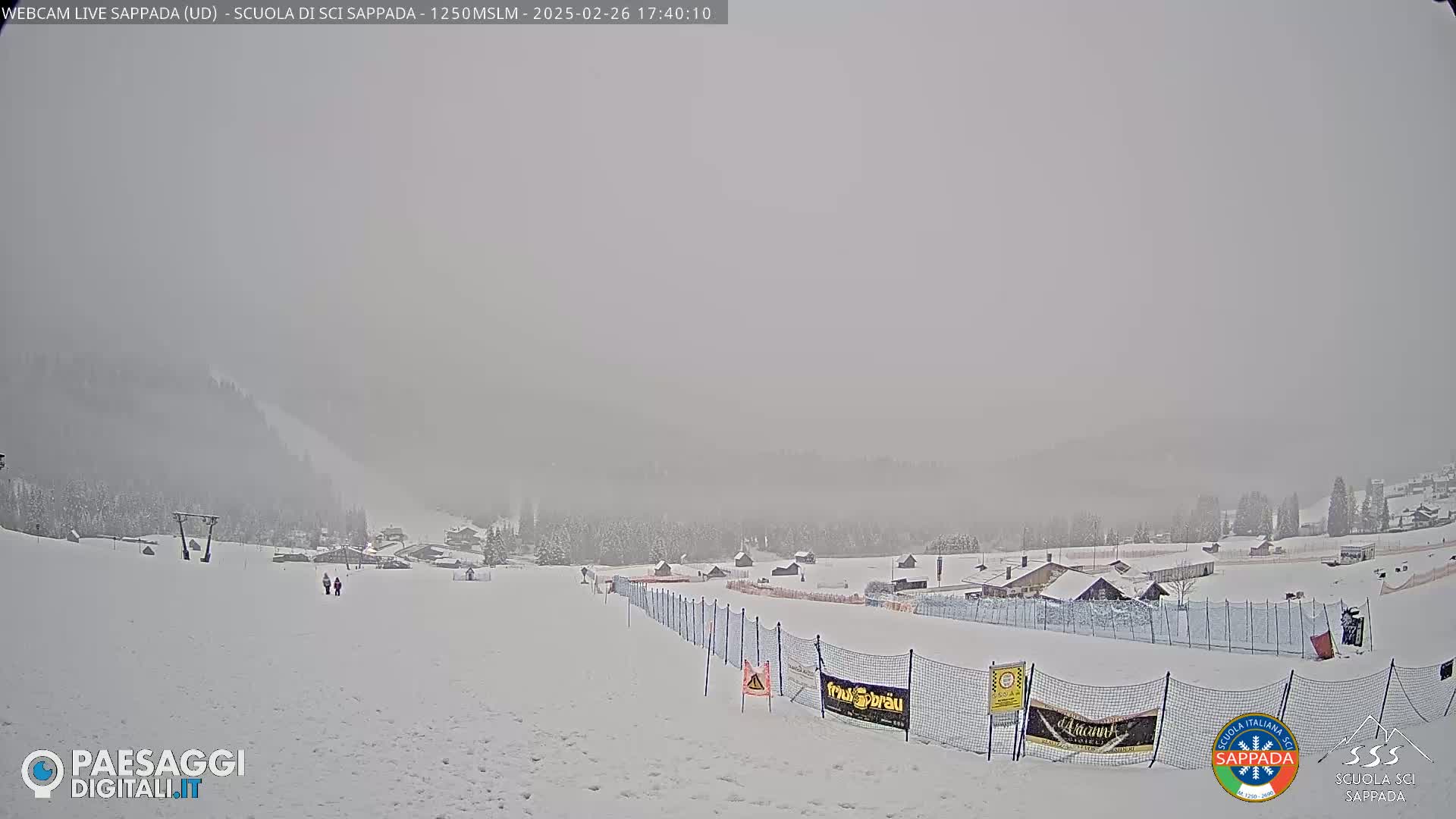 A snowy, foggy scene shows a ski slope with a few people walking, buildings in the distance, and snow-covered trees on the surrounding hills.