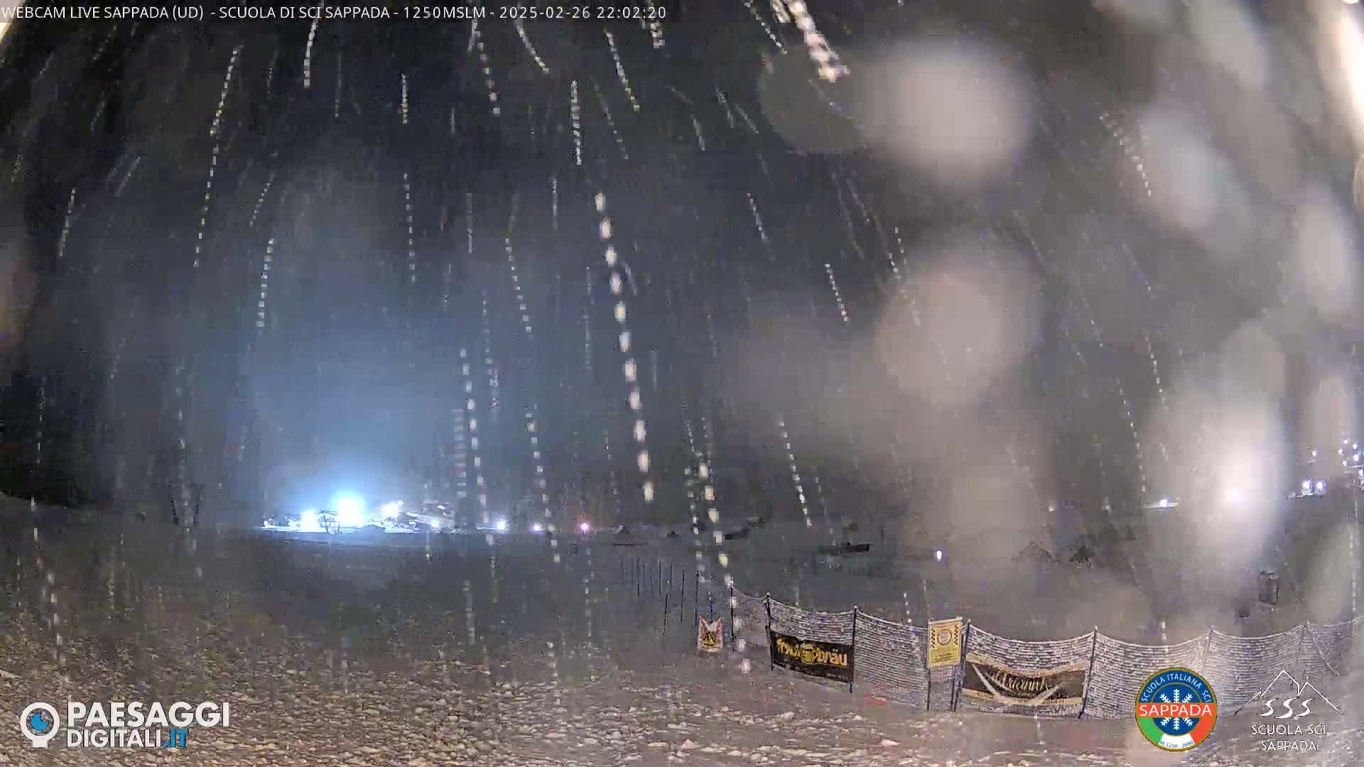 A nighttime snowy scene shows a snow-covered area with lights in the distance, under heavy snowfall.