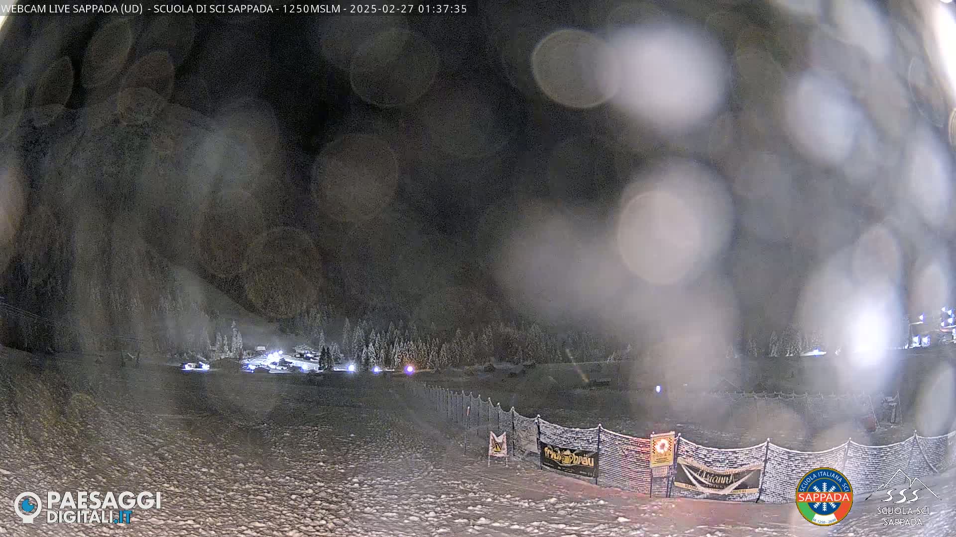 A nighttime snow-covered scene shows a snowy slope with a fence and some lights in the distance, under a dark, foggy sky.