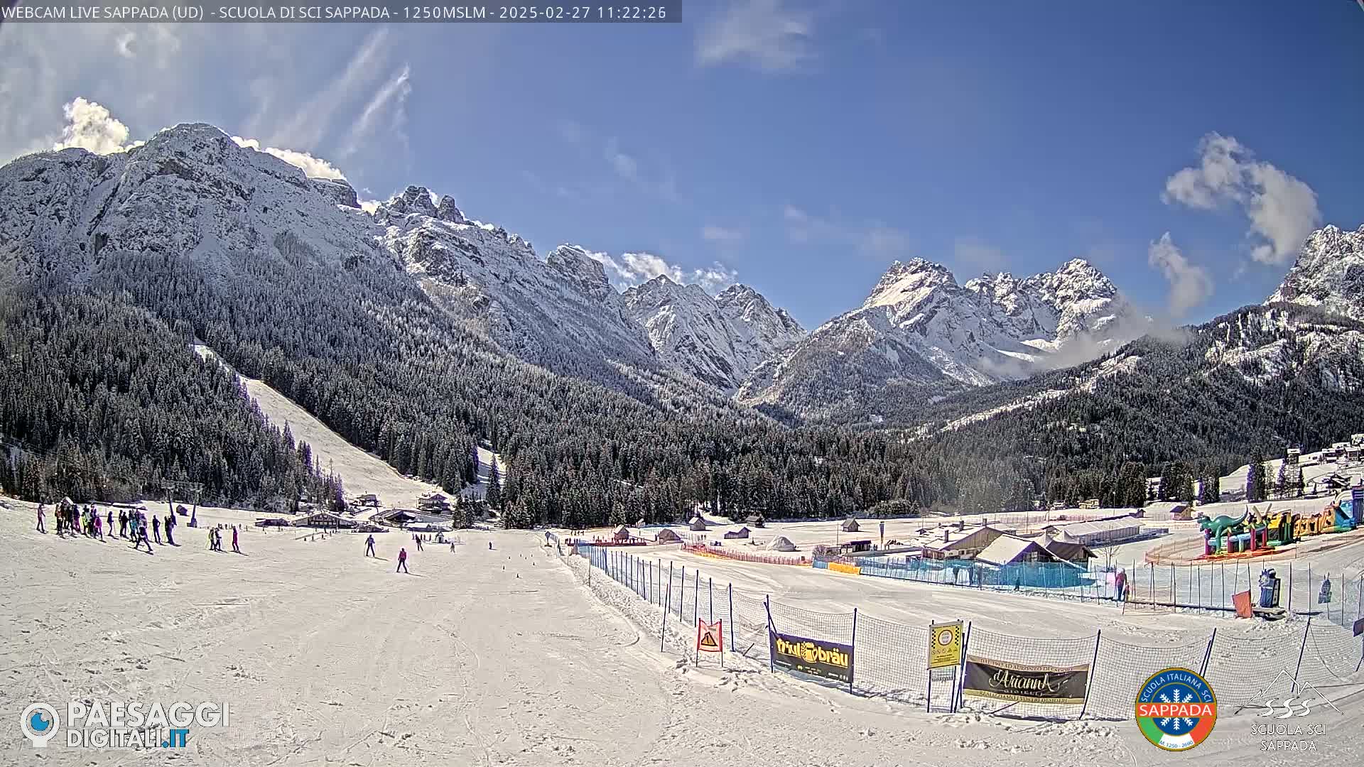 A sunny day at a snow-covered ski resort in the mountains, with several skiers on the slopes and snow-capped peaks in the background.