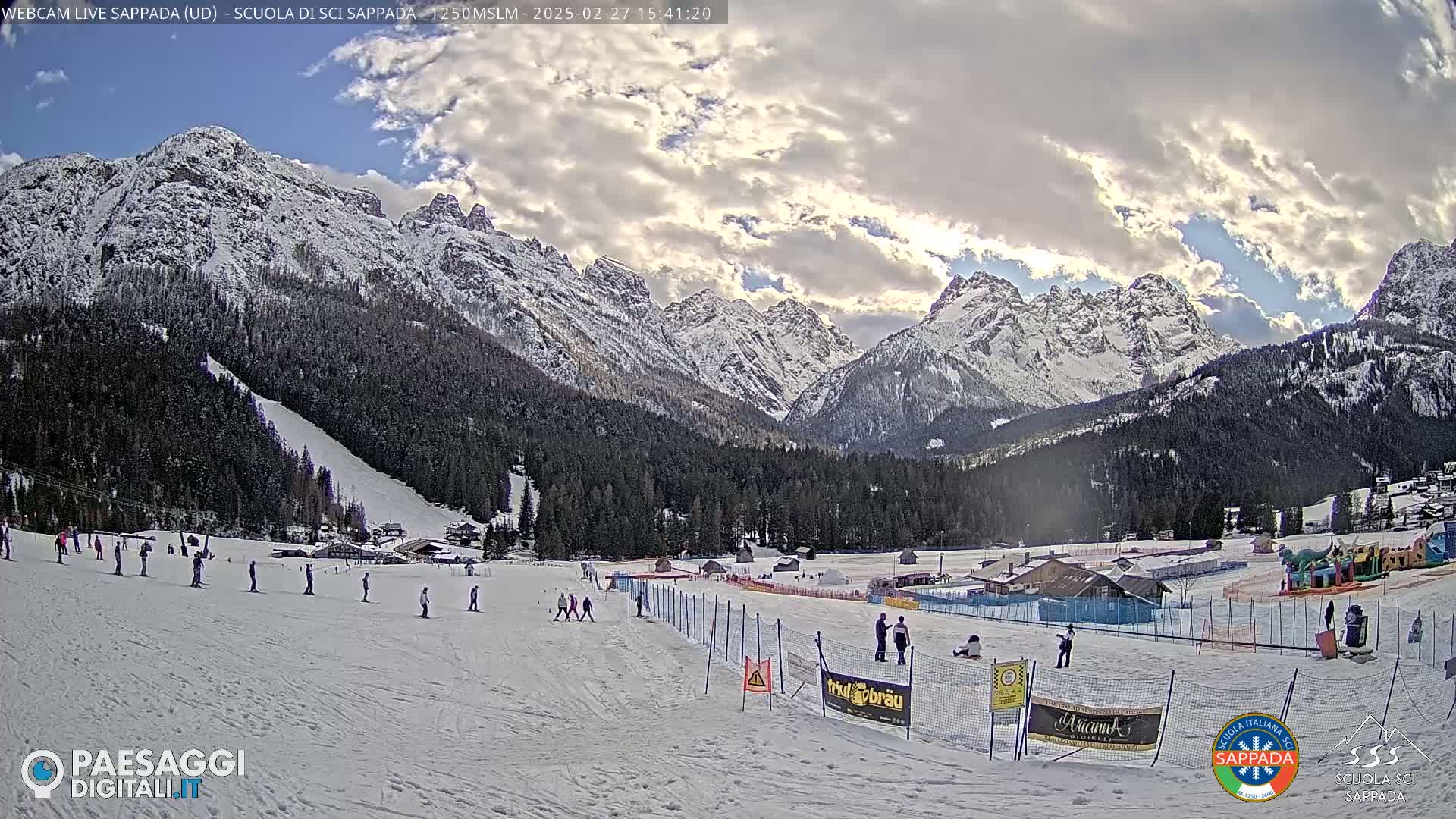 A snow-covered ski slope with several skiers and a backdrop of snow-capped mountains under a partly cloudy sky.