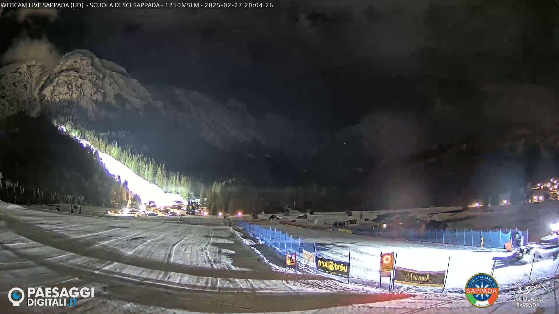 A brightly lit ski slope at night, under a dark, cloudy sky, is situated at the base of a snow-covered mountain.