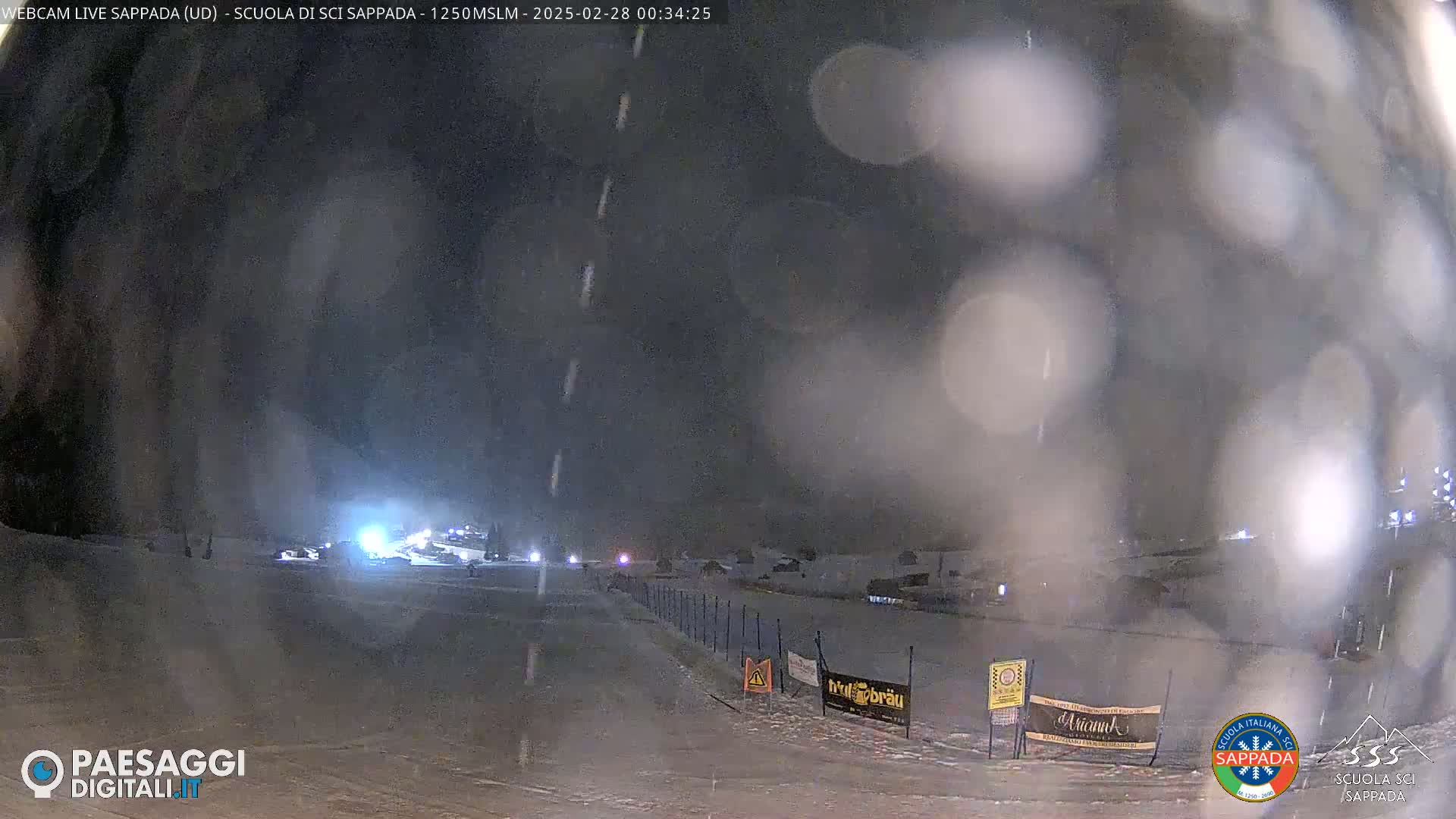 A nighttime view of a snow-covered area with some lights and vehicles visible in the distance, under light snowfall.