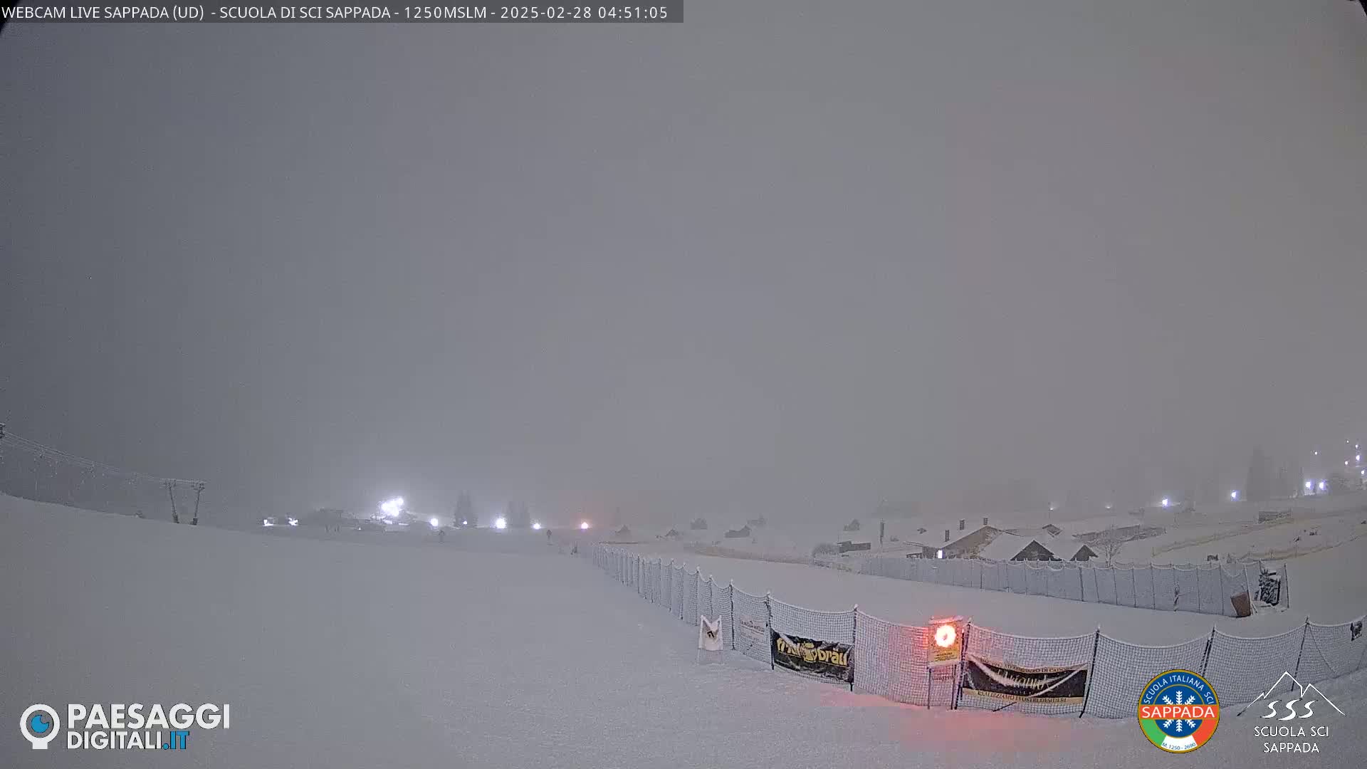 A snow-covered ski slope is visible under heavy snowfall at night, with lights illuminating buildings in the distance.