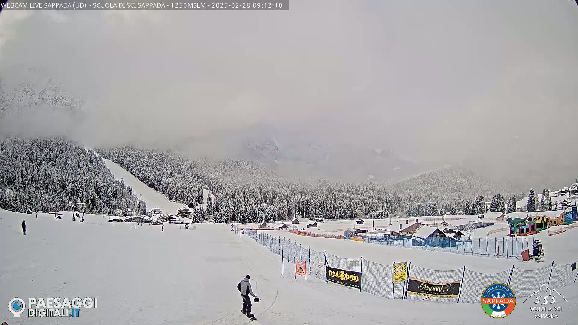 A snow-covered ski slope with a few skiers, surrounded by snow-covered trees and mountains under a foggy sky.