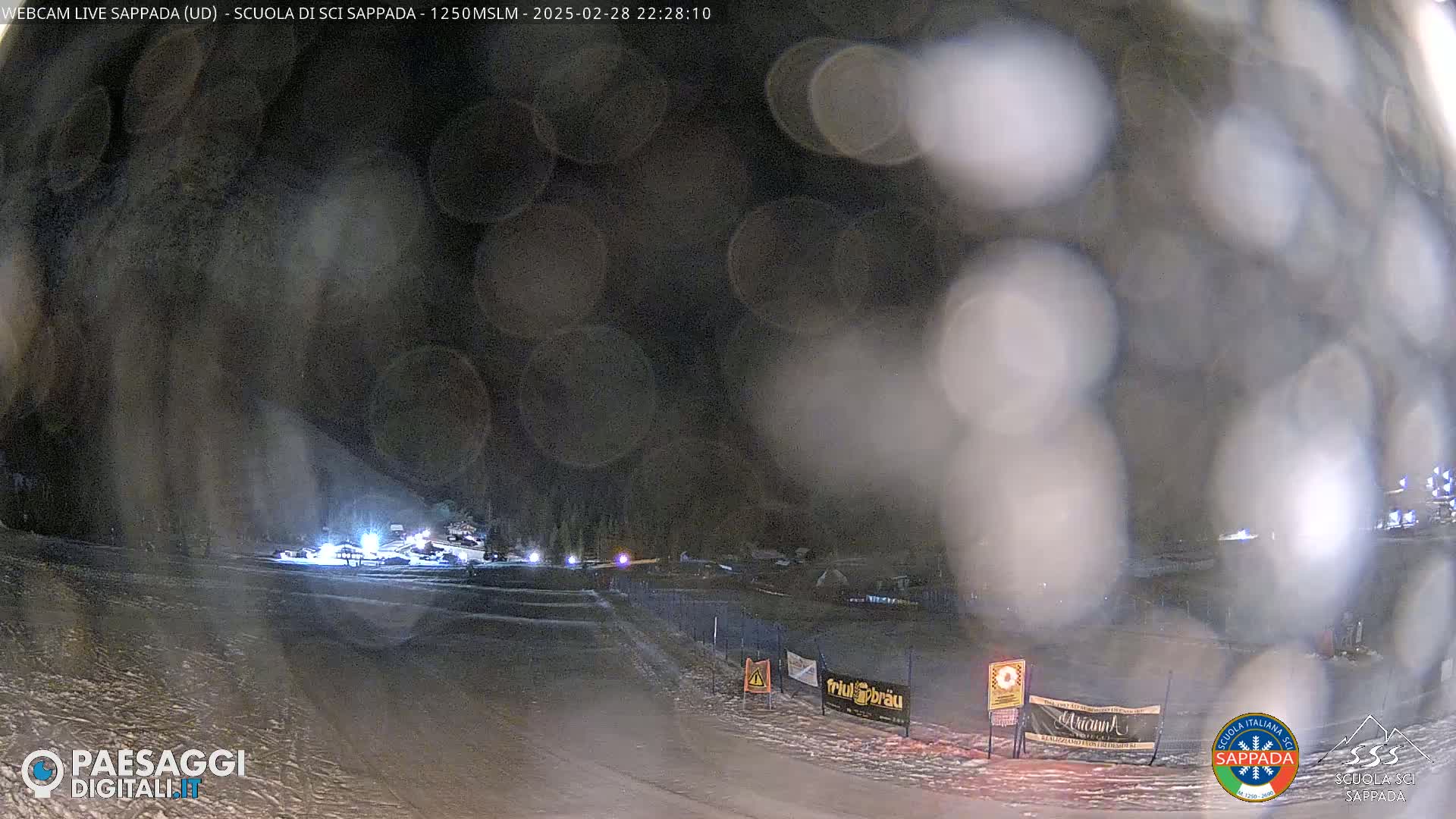 A nighttime view of a snow-covered ski area with several vehicles and lights visible through a blurry, snow-filled lens.