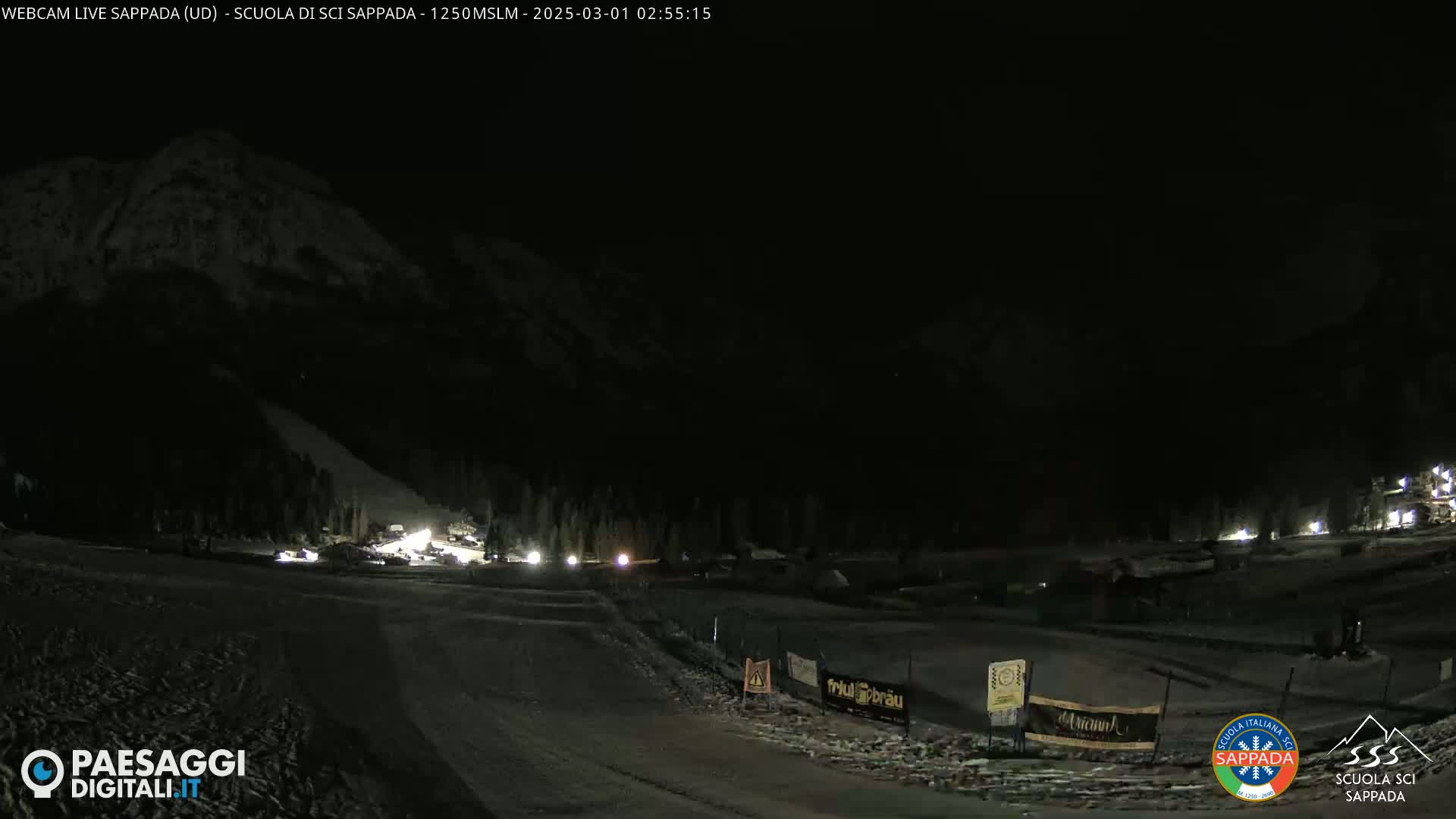 A nighttime, snow-covered ski slope is illuminated by lights, with a dark mountain in the background.