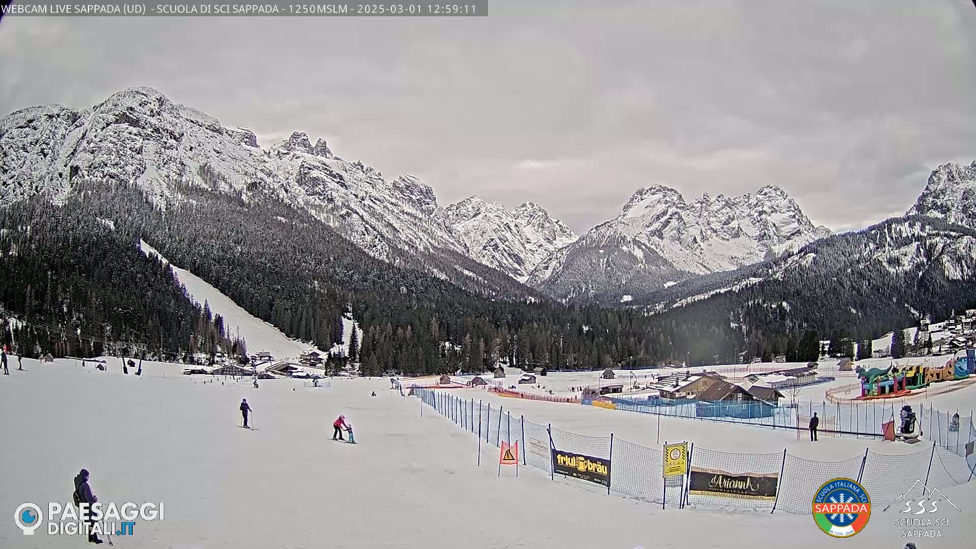 A snowy mountain range is visible in the background, overlooking a snow-covered ski slope with a few skiers and a children's play area.  The sky is overcast.