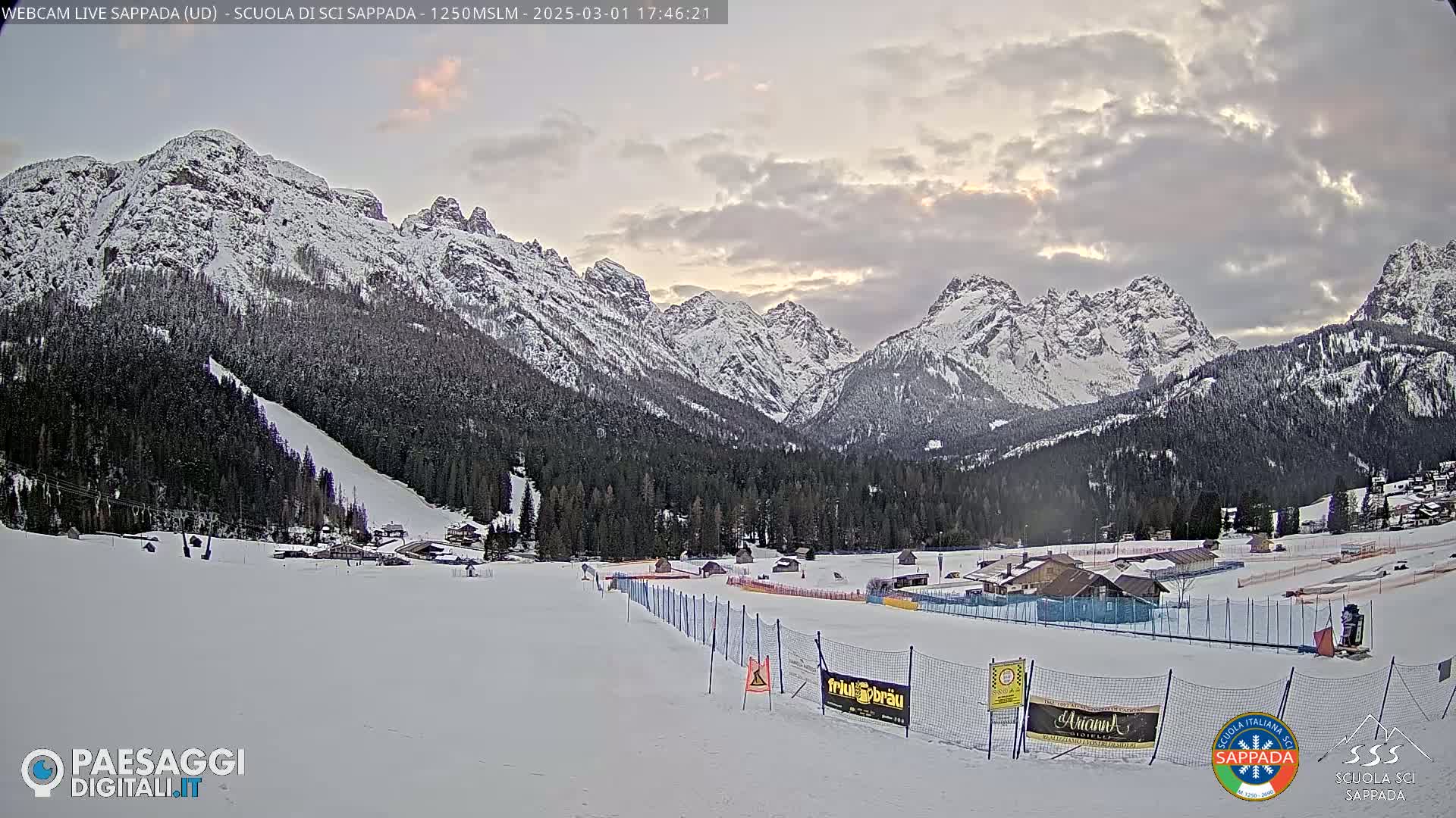 A snow-covered ski resort nestled in a valley between snow-capped mountains under a cloudy sky.