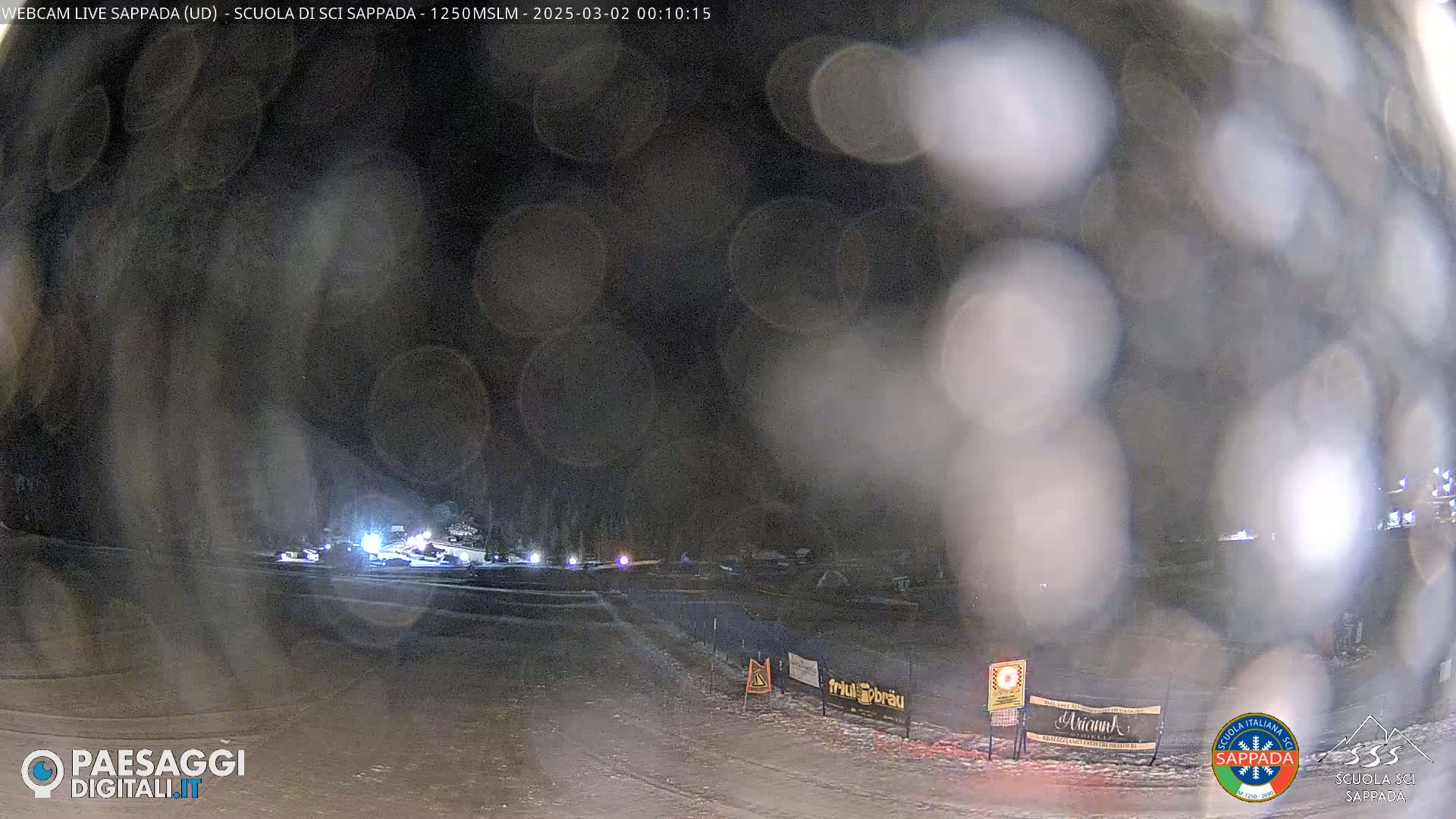 A nighttime, snowy scene shows a mostly empty road or path with a few lights visible in the distance, obscured by blurry, light-colored snow or rain.