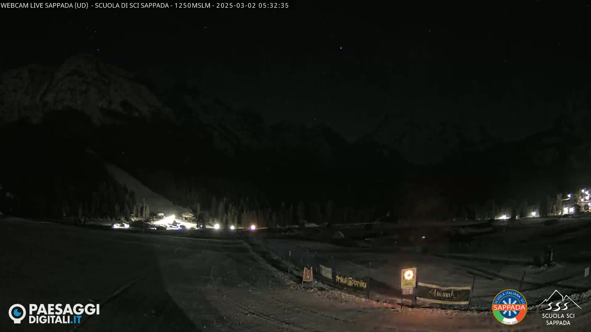 A nighttime view of a snow-covered mountain slope with a few lights visible in the distance under a clear, starry sky.