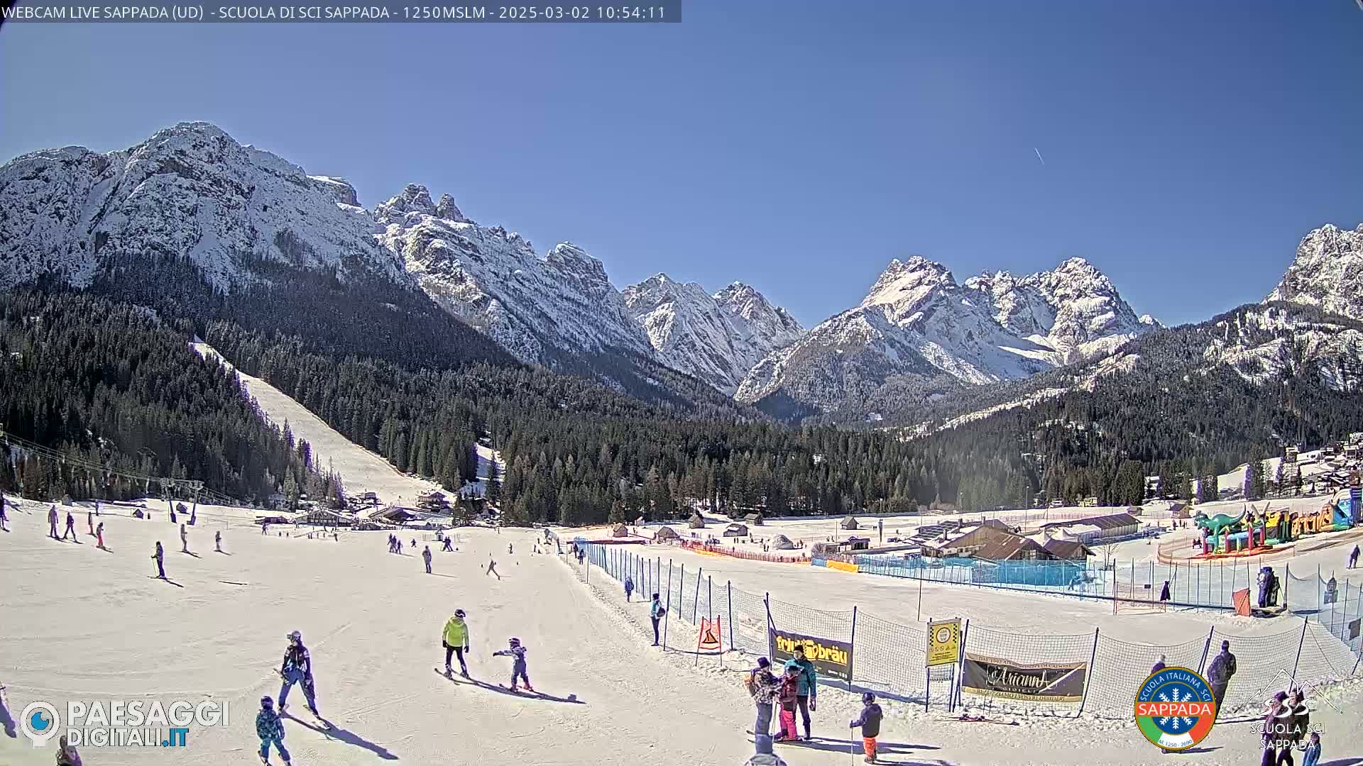 A sunny day at a snow-covered ski resort in the mountains, with skiers and snowboarders on the slopes.