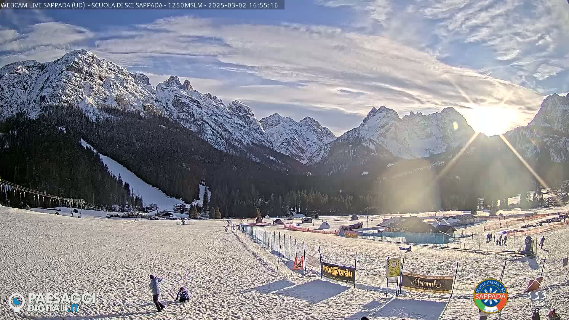 A snow-covered ski slope sits below sunlit, snow-capped mountains under a partly cloudy sky.