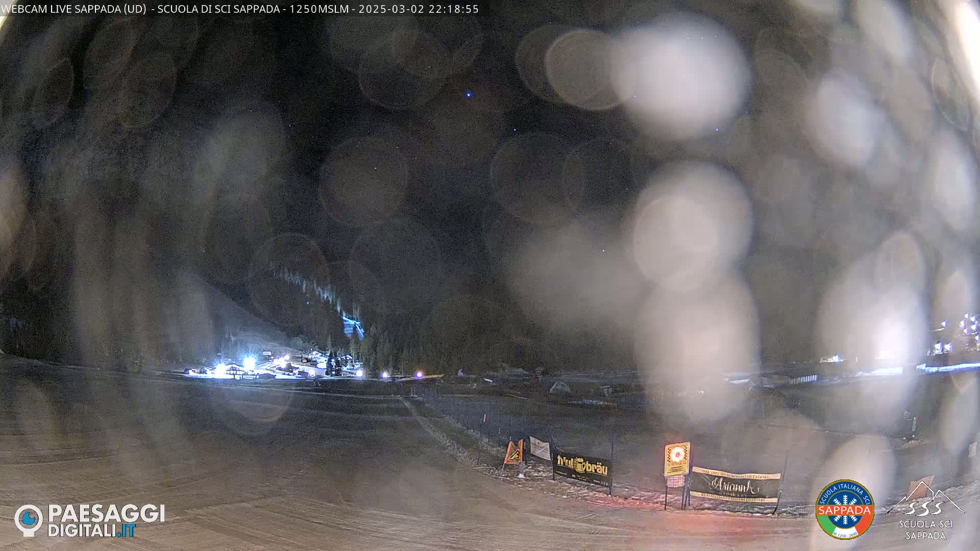 A nighttime view of a snow-covered ski slope with some lights visible in the distance, under a dark, slightly hazy sky with a few stars.