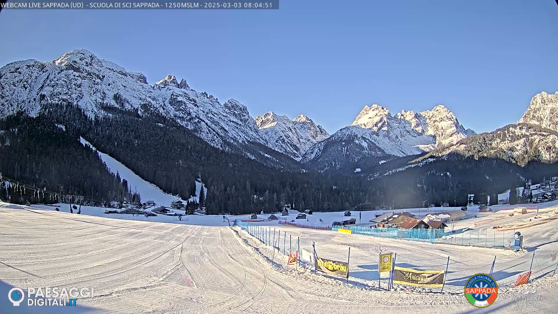 A snow-covered ski slope is situated in a valley, with snow-capped mountains and a clear blue sky in the background.