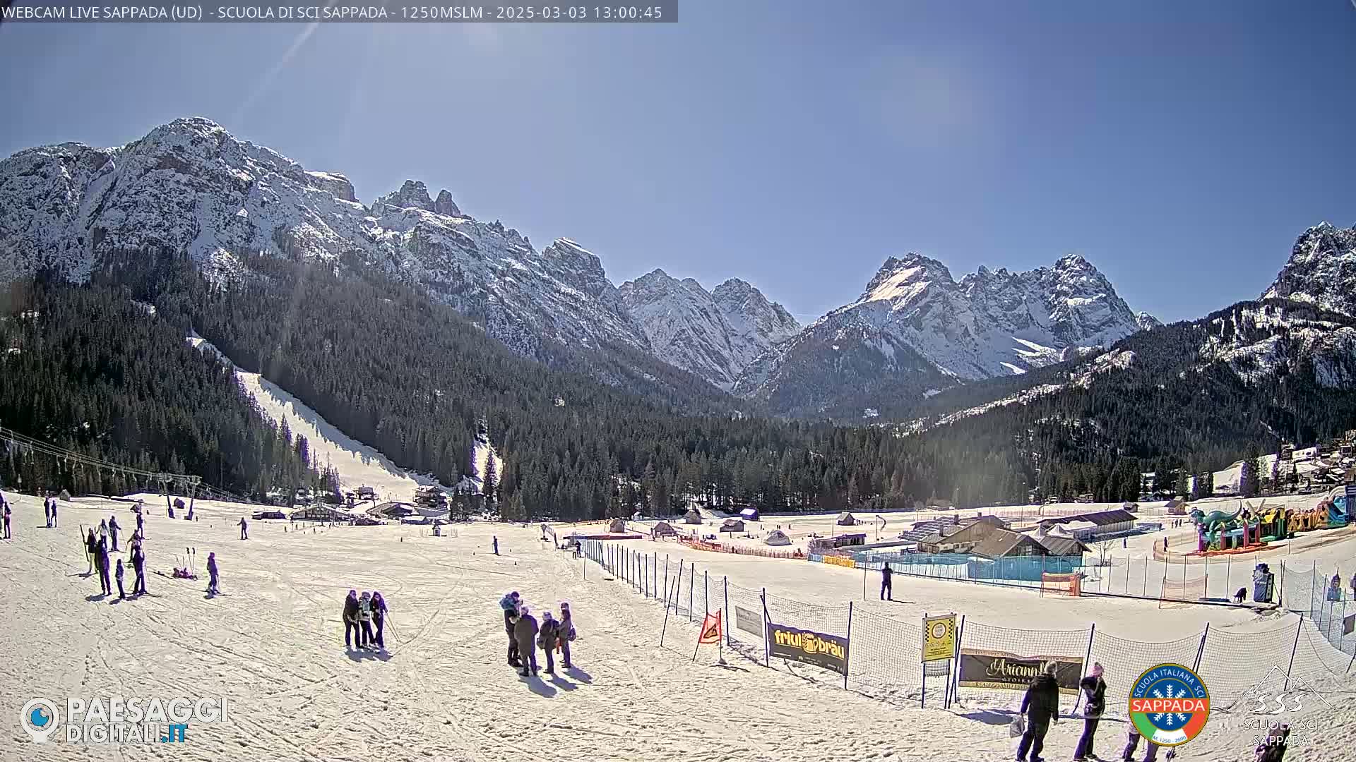 A sunny day at a snow-covered ski resort in the mountains, with skiers and people standing around on the slopes and snow-capped peaks in the background.
