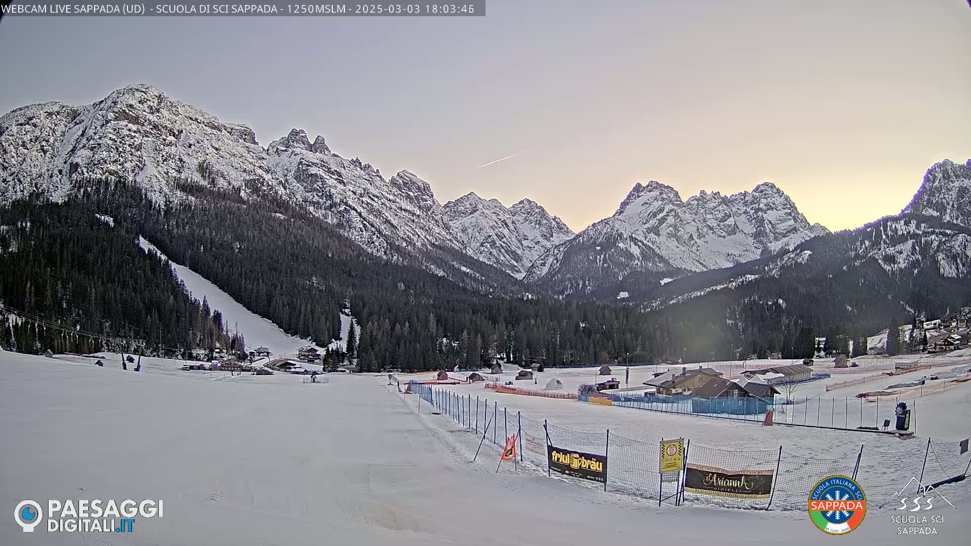 A snow-covered ski resort nestled in a valley between snow-capped mountains under a pale twilight sky.