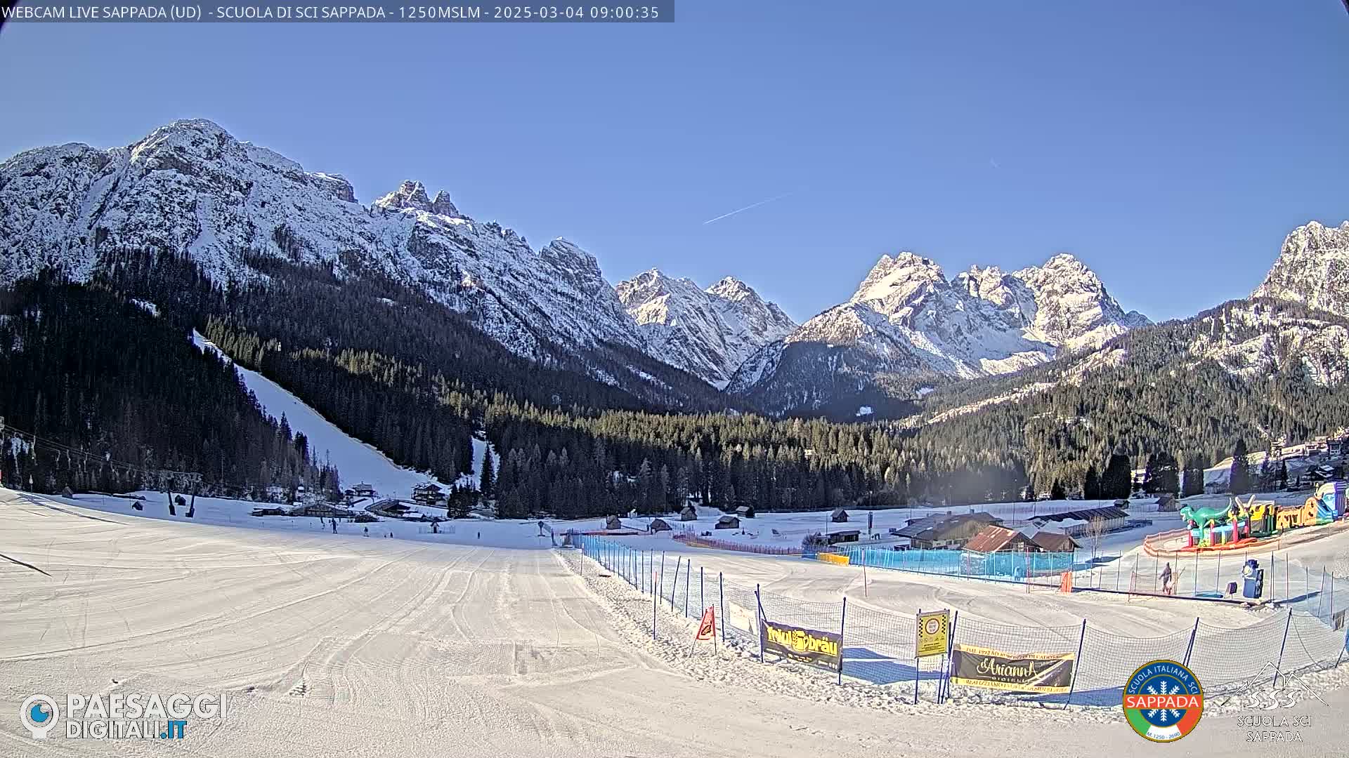 A snow-covered ski slope at the base of snow-capped mountains under a clear blue sky.