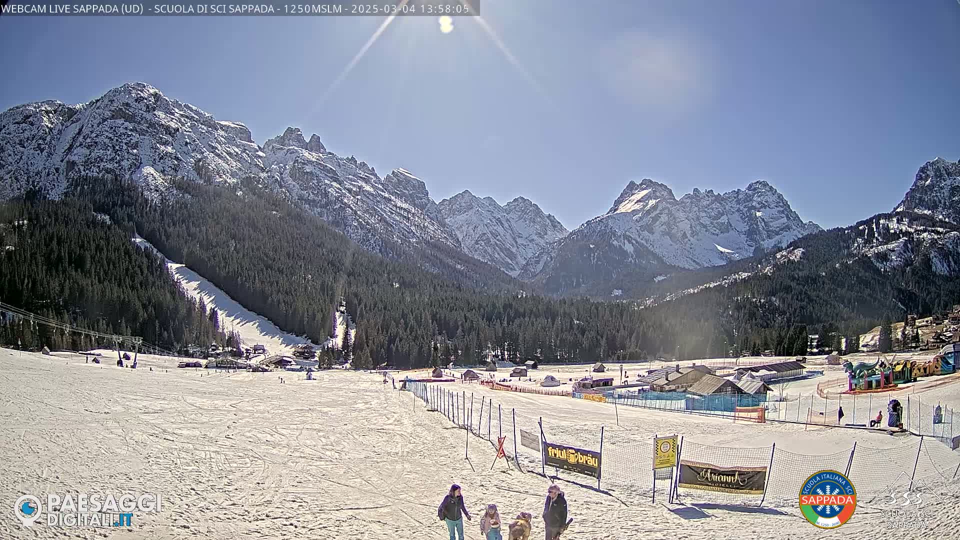A snow-covered ski resort nestled in a valley, with snow-capped mountains in the background under a clear, sunny sky.
