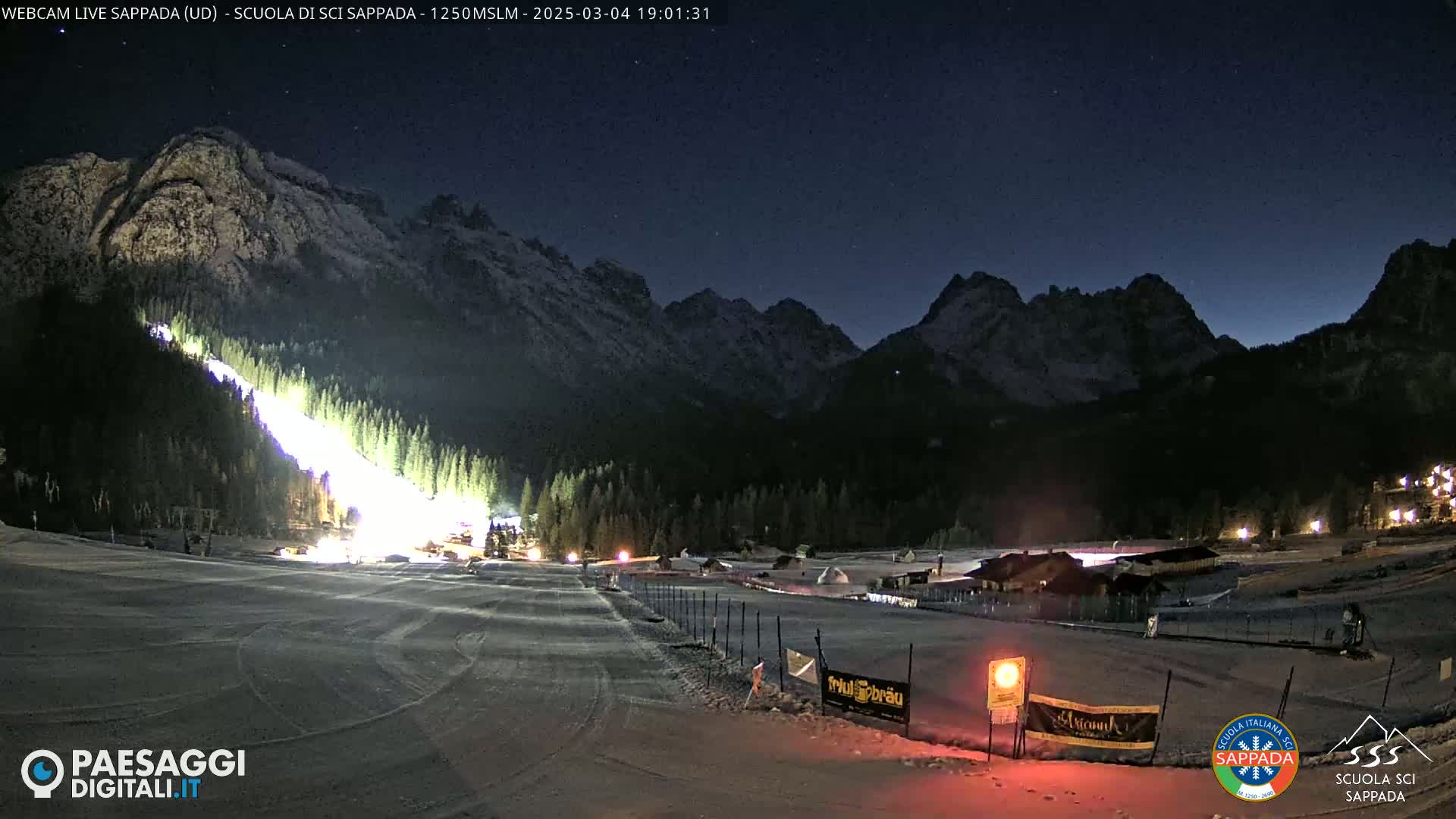 A brightly lit ski slope at night, surrounded by snow-covered mountains under a dark, clear sky.