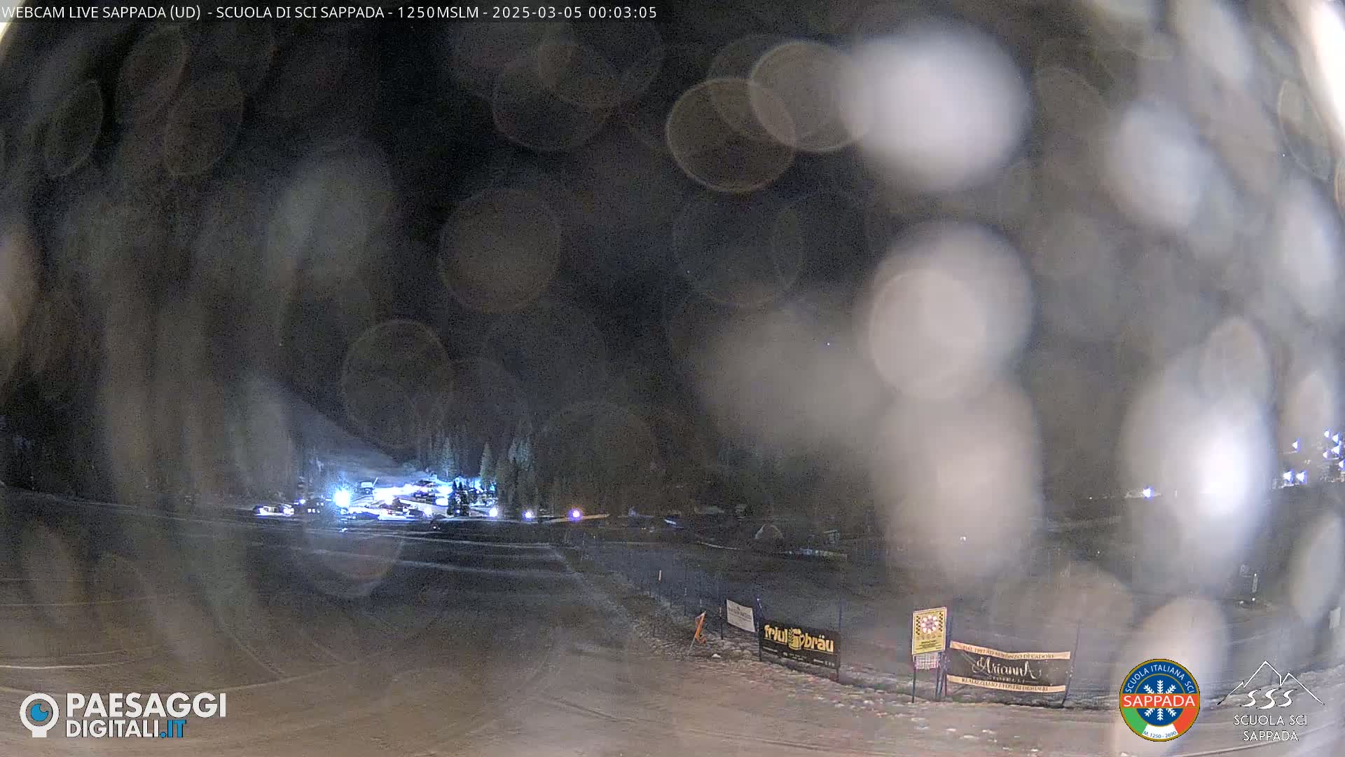 A nighttime view of a snow-covered area with some vehicles and lights visible through blurry, possibly rain- or snow-covered glass.
