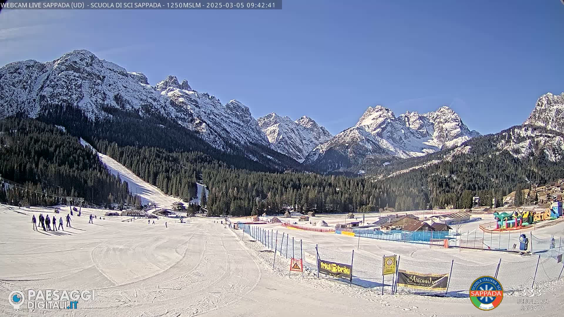 A snow-covered ski slope with several people skiing and a backdrop of snow-capped mountains under a clear blue sky.