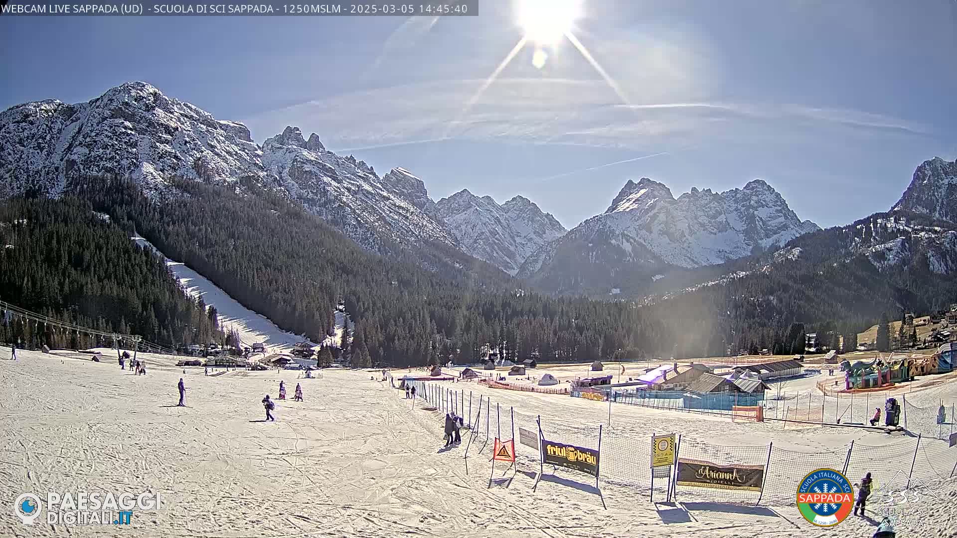 A sunny day at a snow-covered ski resort in the mountains, with several skiers visible on the slopes.