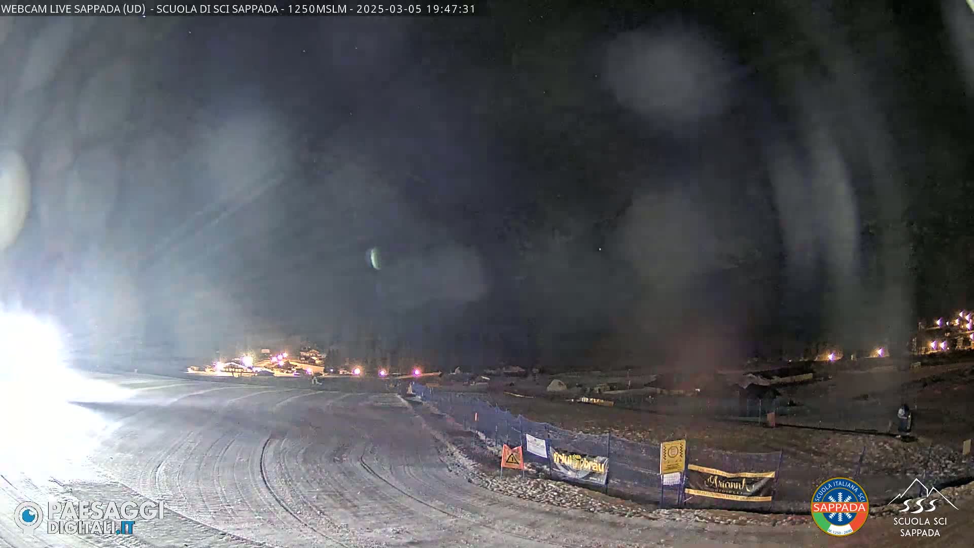A nighttime view of a snow-covered ski slope with some lights visible in the distance under a cloudy, dark sky.