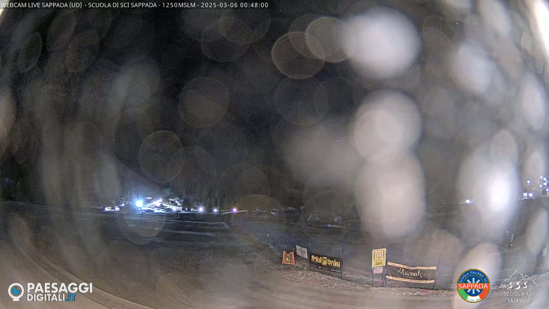 A nighttime view of a snow-covered area with several vehicles and blurry lights in the distance, under a dark, possibly hazy sky.