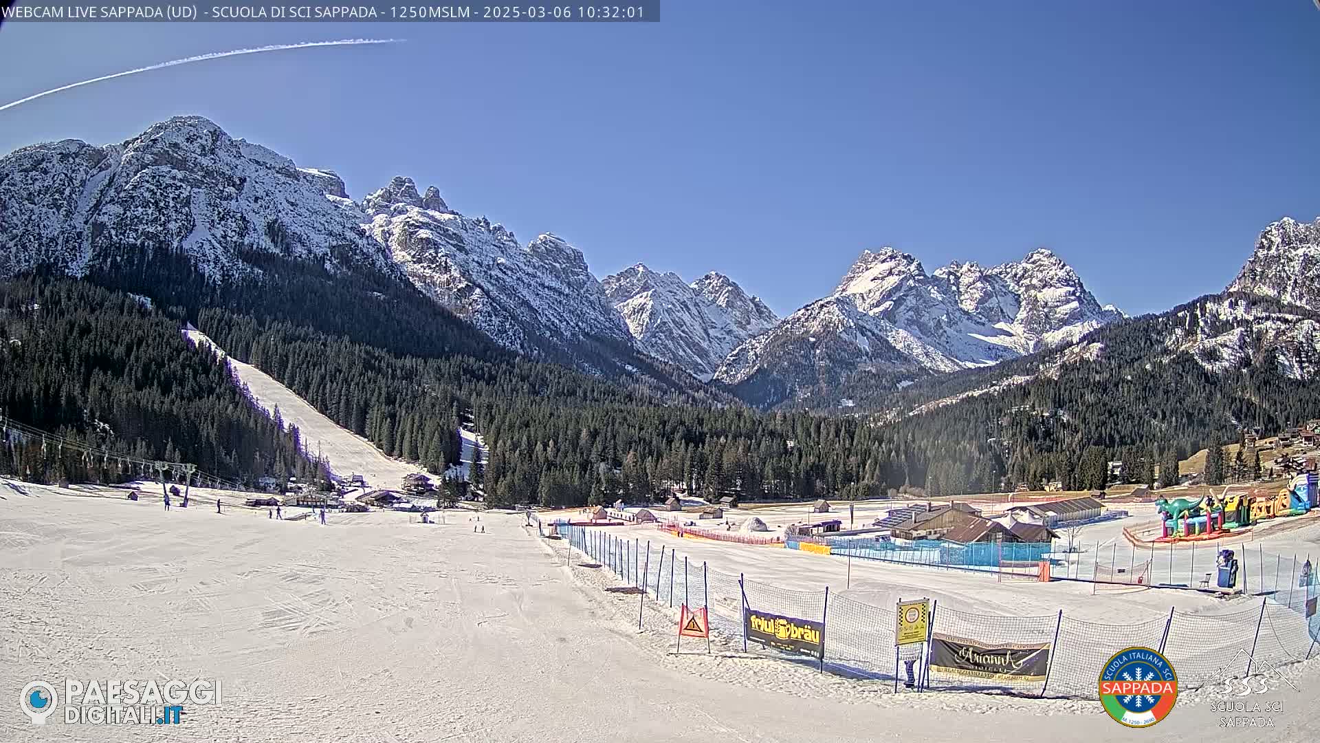 A snow-covered ski slope at the base of snow-capped mountains under a clear blue sky.