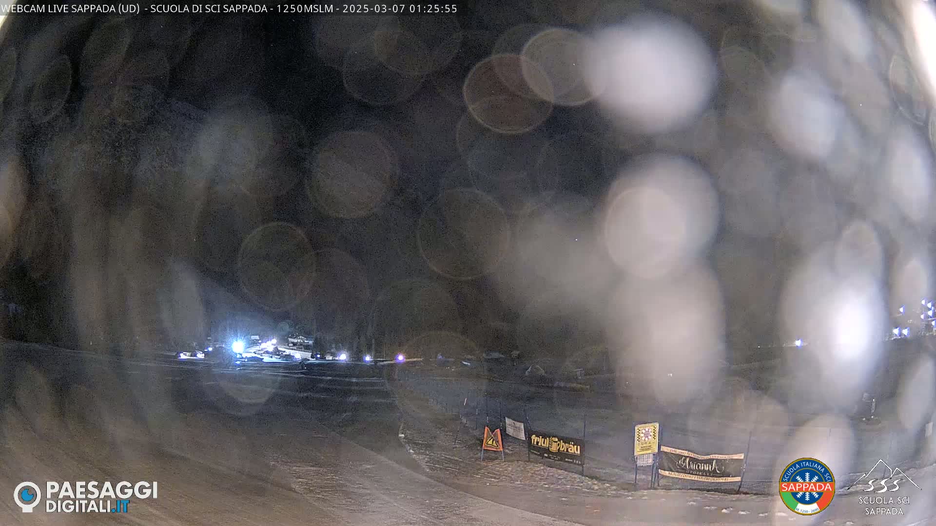 A nighttime, snow-covered scene shows a road with several vehicles, blurred by a layer of what appears to be condensation or light snow on the camera lens.